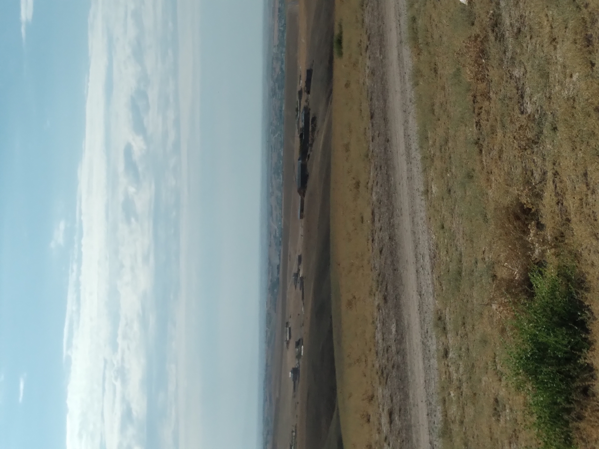 The image depicts a dry, arid landscape with rolling hills under a cloudy sky. In the mid-ground, several low-lying buildings, possibly farm structures or rural dwellings, are scattered across the terrain. A dirt road runs across the foreground, cutting through dry, patchy vegetation. In the far distance, a larger settlement or town is visible, appearing as a cluster of buildings on the horizon. The overall impression is of a rural or semi-arid environment, consistent with the outskirts of a town like Aktepa, Uzbekistan, characterized by open land and dispersed human settlements.