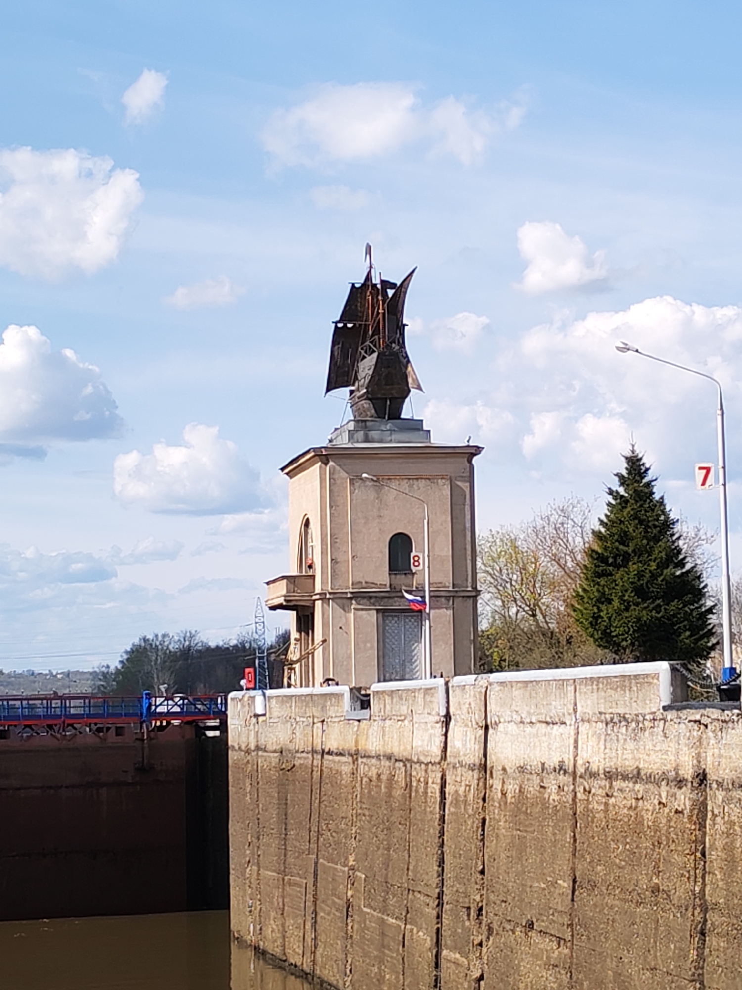 The image depicts a lock on the Moscow Canal, located in the city of Yakhroma, Russia. A tower with a metal sculpture of a ship on top stands next to the lock. The tower is painted a beige color and has arched windows. A small balcony is visible on one side of the tower. Numbered signs "7" and "8" are attached to nearby poles. A green evergreen tree is situated next to the tower, and a street light is also present. The lock itself is constructed from large, weathered concrete blocks. A blue and red bridge is visible in the background. The sky is blue with scattered white clouds.