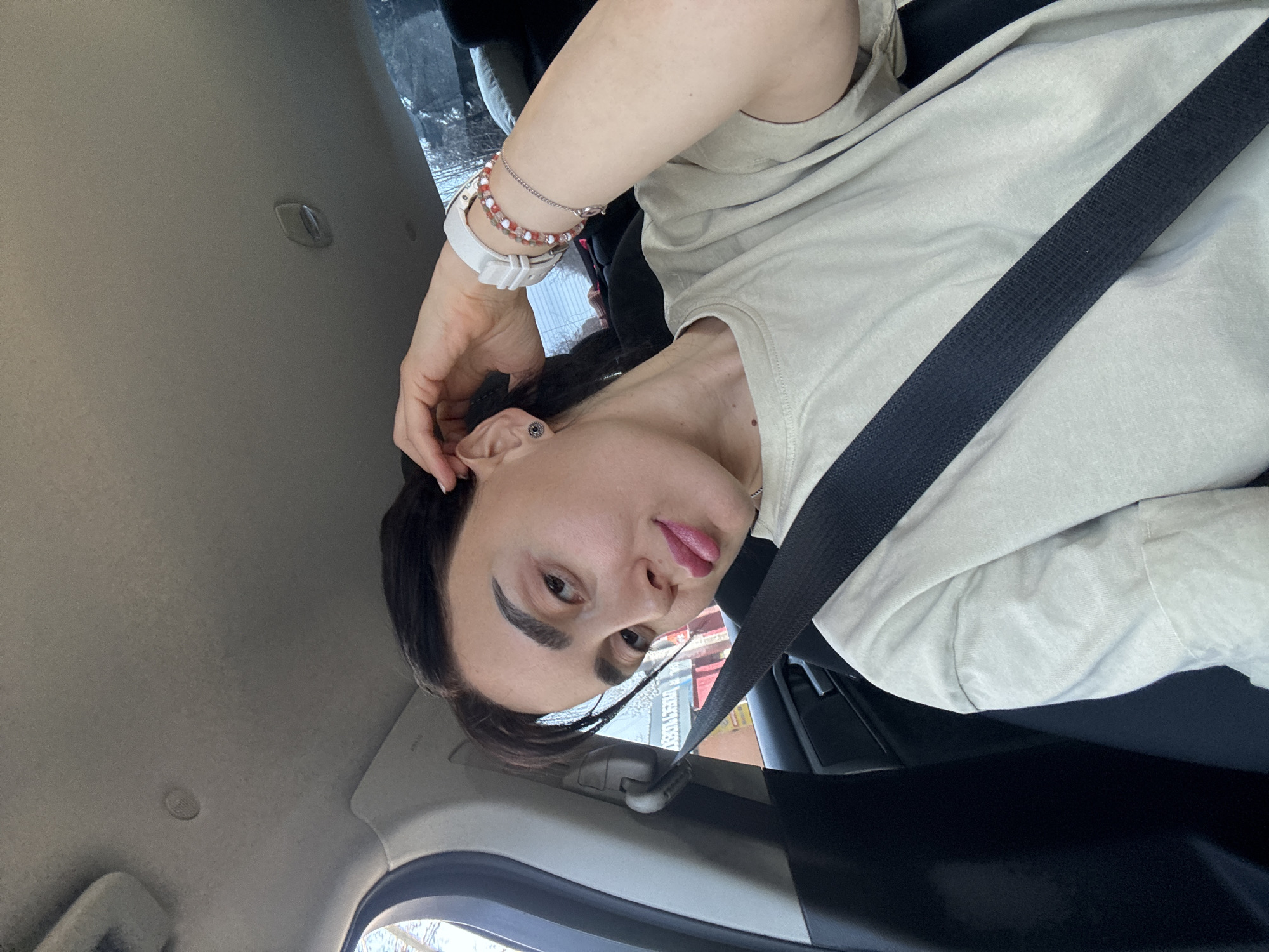 A young adult woman is inside a vehicle, wearing a seatbelt and a beige-colored t-shirt. She has dark hair and is wearing a wristwatch and bracelets. The background visible through the vehicle's window suggests an urban environment in Tbilisi, Georgia, based on the architectural details.