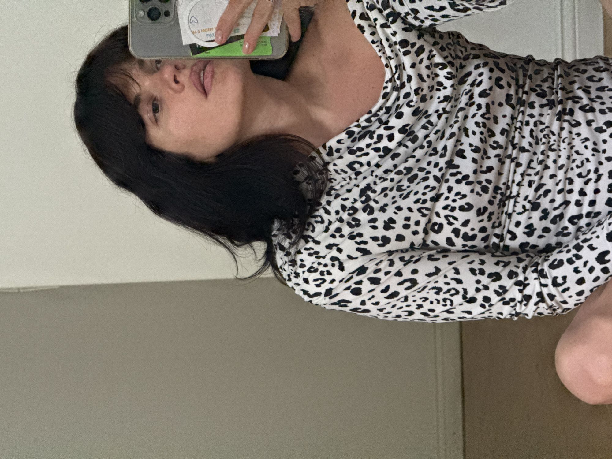 A woman with dark hair is taking a selfie. She is wearing a white and black patterned long-sleeved shirt. She is holding a silver smartphone in front of her face. The background appears to be an interior wall and a door. There is no specific information to identify the location as Berlin, Germany.