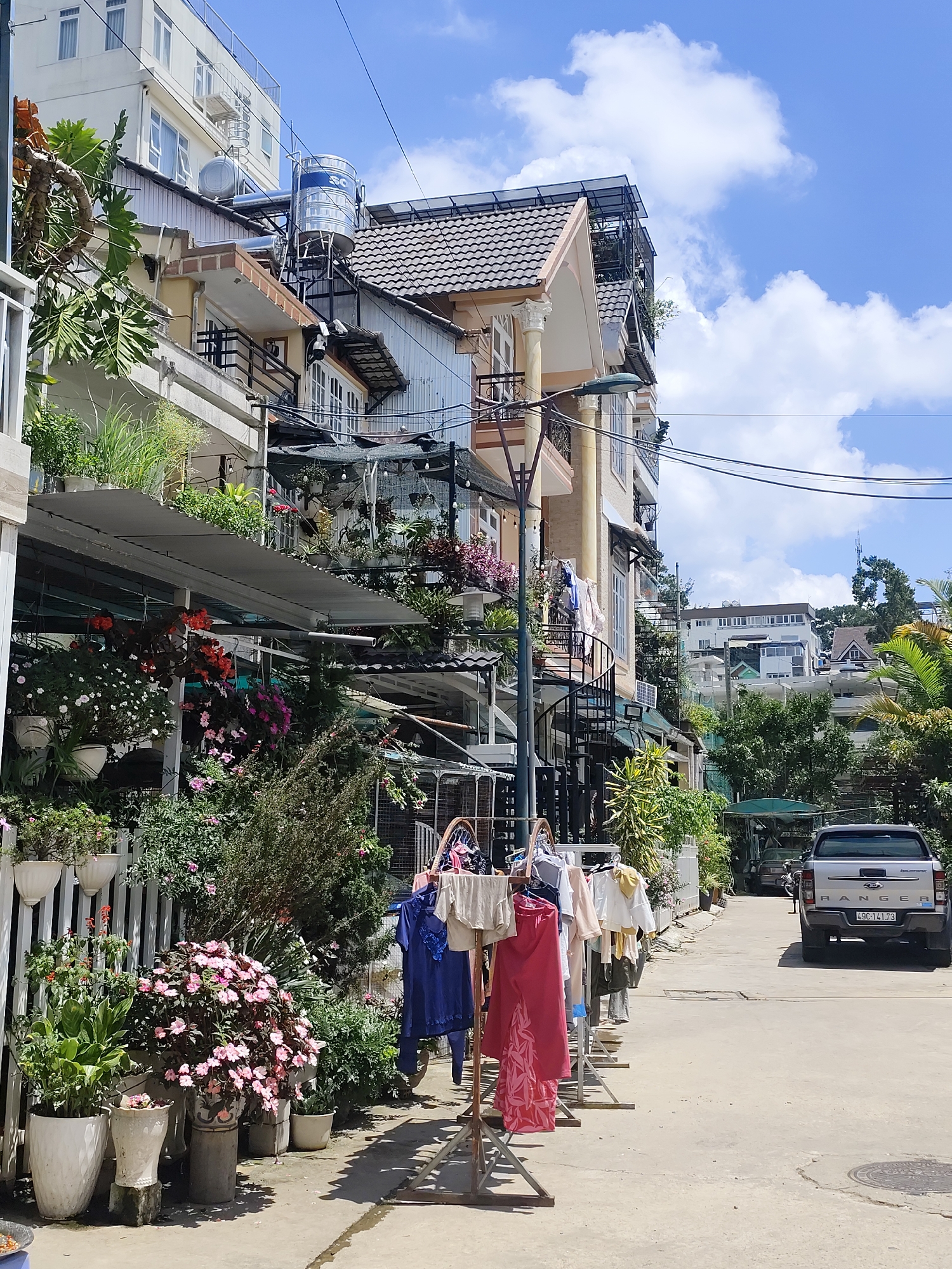 This is a medium shot of a residential street in Da Lat, Vietnam. The buildings are multi-story with balconies adorned with numerous potted plants. Clothes are hung on a rack in the foreground, with various garments including a blue dress and a pink skirt. A white pickup truck is parked further down the street. The sky is blue with scattered white clouds. The scene depicts a typical urban environment in Da Lat, known for its lush greenery and often decorated balconies.