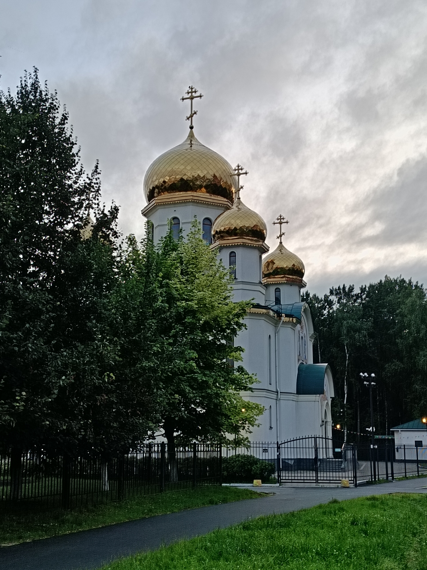 An exterior, eye-level, full shot captures a white orthodox church with golden domes, situated in a naturalistic urban setting. The church's architecture is characterized by its multiple domes, each topped with a cross. A dark, overcast sky looms above, with visible clouds. In the foreground, a paved pathway runs diagonally from the bottom left corner towards the center of the image, bordered by a strip of green grass. A black metal fence runs horizontally across the mid-ground, separating the pathway and grassy area from the church grounds. Behind the fence, lush green trees and foliage partially obscure the church. The church itself is a prominent structure with arched windows and decorative architectural elements. A large black metal gate with decorative scrollwork is visible in front of the church entrance. Beyond the church, dense green trees form a backdrop. The lighting suggests it is late afternoon or evening, with diffused light contributing to the subdued atmosphere. No people are visible in the frame.