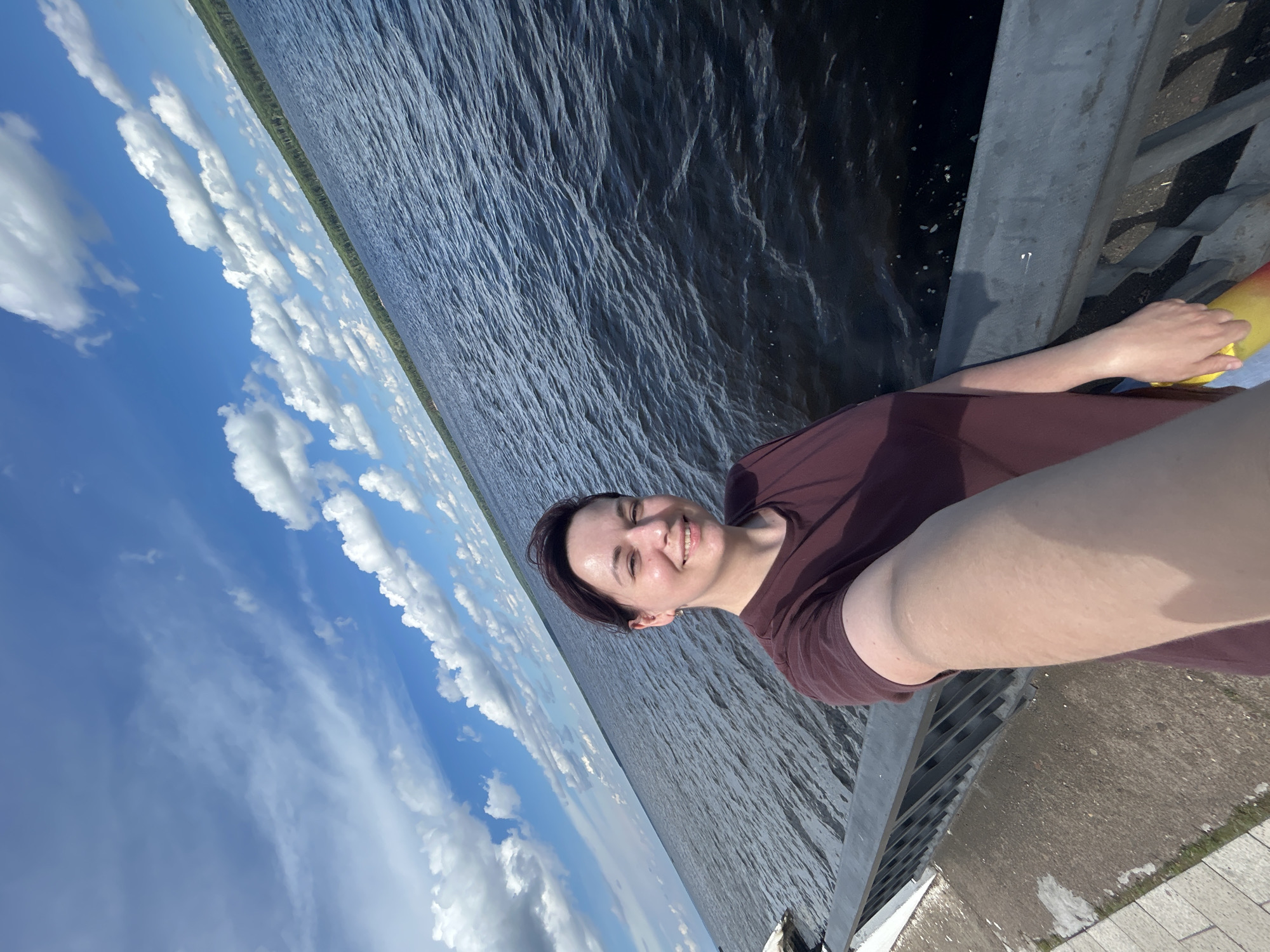 In Chaikovsky, Russia, a young woman with short, dark hair and a maroon t-shirt takes a selfie outdoors. She is standing on a walkway with a railing, next to a large body of dark water. The water has small ripples, suggesting it is windy. The sky above is blue with scattered white clouds, and a line of green trees is visible in the distance along the horizon. The woman is smiling and holding a yellow and red object, possibly a bottle or a bag, in her right hand. The walkway appears to be paved with light-colored stones or tiles.