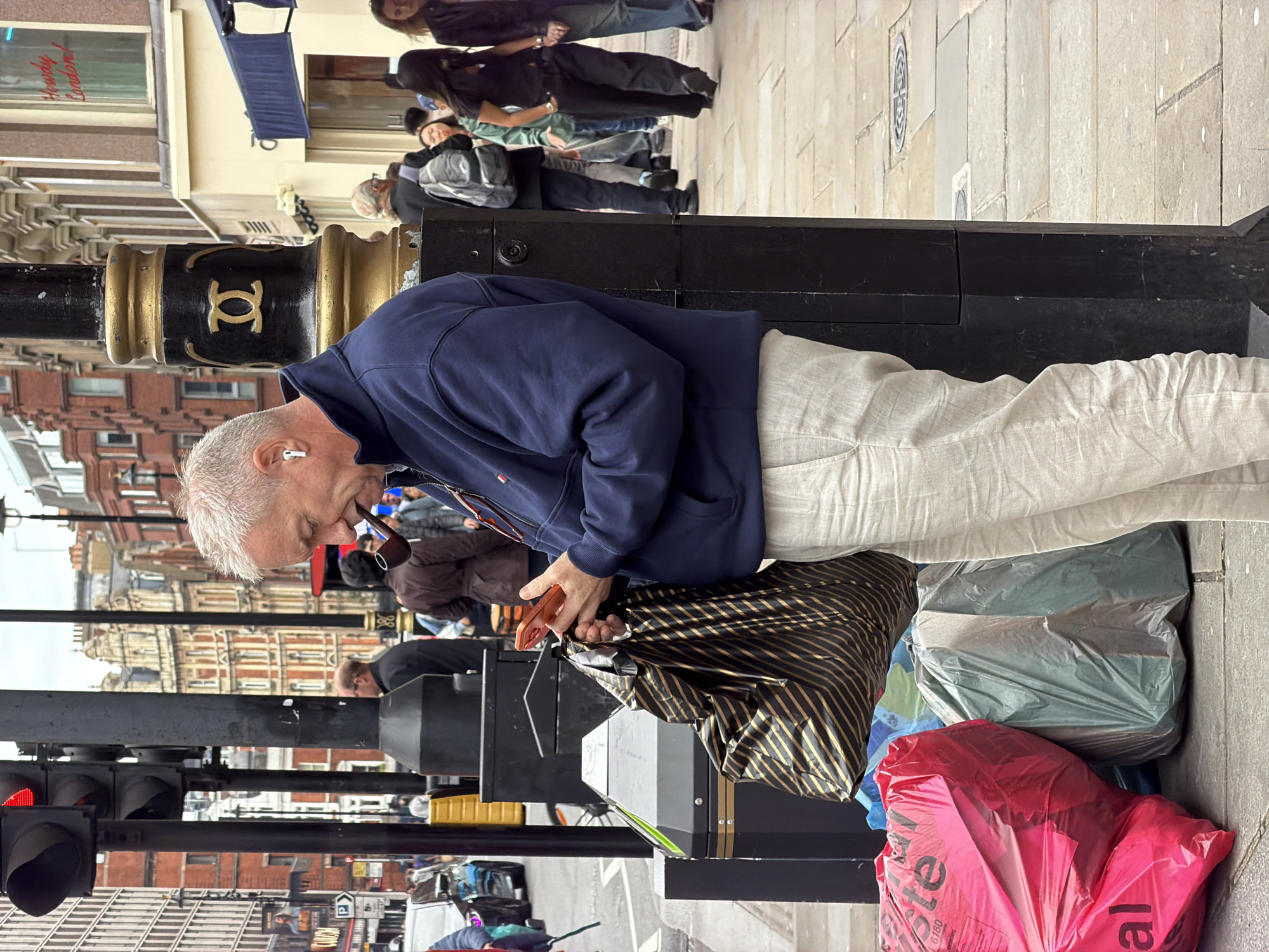 The image depicts a daytime urban street scene in London, United Kingdom, characterized by pedestrian activity and distinct architecture.
In the foreground, a light-skinned man with short, spiky grey hair stands facing slightly left. He wears a navy blue zippered jacket, featuring a small red, white, and blue logo on the left chest, and light-colored, possibly linen, trousers. A white wireless earbud is visible in his right ear. He holds an orange-cased smartphone in both hands, looking down at the screen. A dark brown pipe is held in his mouth but not actively being smoked. A large, striped black and gold/tan reusable shopping bag hangs from his left hand, resting on the black street furniture next to him.
To the man's immediate left, at the base of the street furniture, are several discarded large plastic bags: a prominent bright red one marked "Waste 6180" and "al" (partially visible), and two greyish-green ones.
The man stands next to a robust black street pole, which features an ornate gold band with a decorative symbol (two interlocking 'C's or similar) and additional gold banding above and below. Below this, the pole transitions into angular black utility boxes or bases.
In the mid-ground, behind the man and pole, a diverse group of pedestrians walks along a wide pavement. A person in a brown jacket stands near another black pole, seemingly holding a dark object to their mouth. Further right, a man with a grey backpack walks past. On the far right, a shop window clearly displays "Howdy London!" in red script.
The background reveals a mix of London architecture. On the left, tall, modern buildings are visible, along with a traffic light showing a red signal and a billboard displaying "CULOM." In the center, an elaborate red brick building with numerous windows and ornate detailing stands out. The street pavement is made of flagstones. The sky is overcast, indicating diffuse lighting without harsh shadows.