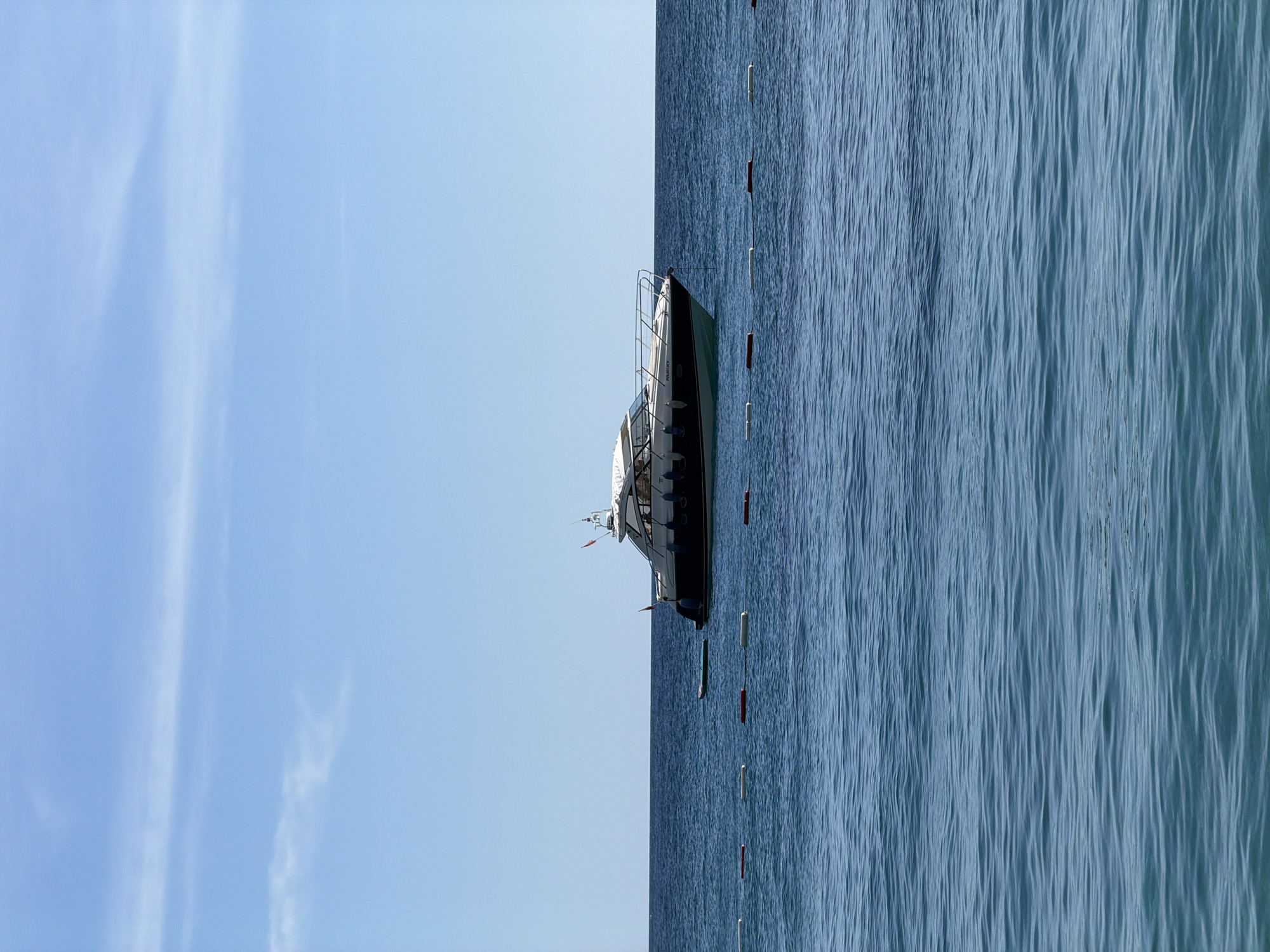 The image presents a boat positioned near a safety barrier in the water, possibly in Sveti Stefan, Montenegro. The sea is visible on the right side of the image, with a pattern of ripples indicating water movement. The sky appears cloudless and bright blue on the left side of the image. The safety barrier is marked by intermittent white and red buoys. The boat, with its sleek design, is a focal point, with masts. The overall scene suggests a calm, clear day at sea, potentially in a designated swimming or boating area.