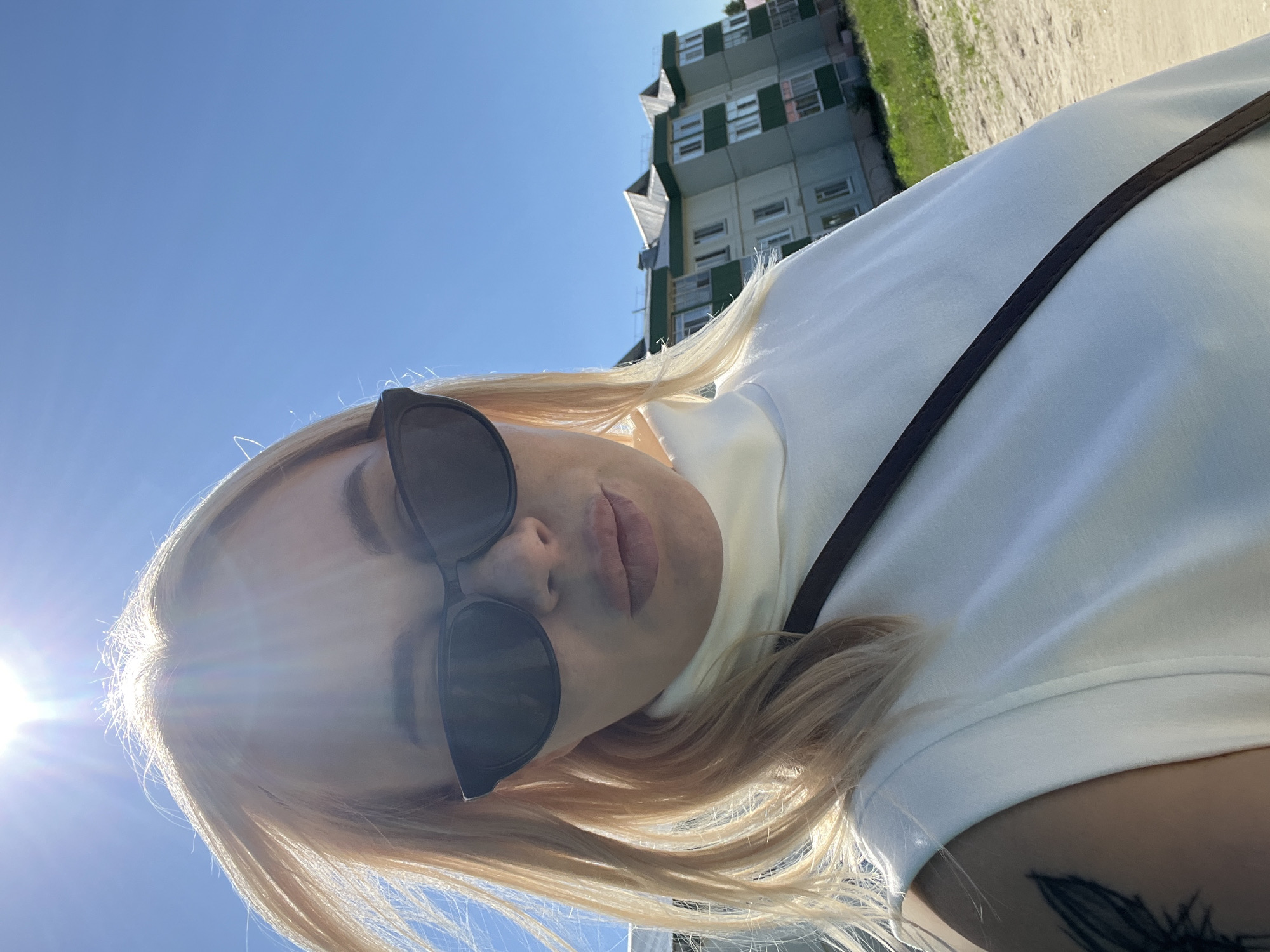 A young woman with blonde hair and sunglasses takes a selfie outdoors on a sunny day. She is wearing a white t-shirt and a dark strap is visible across her chest. Her lips are slightly parted and she is looking towards the camera. Her hair is blowing in the wind and the sun is shining brightly, causing lens flare. In the background, there is a blue sky, a modern building with green and grey panels, and a sandy area with some green grass. The image is rotated, with the woman appearing upside down. The overall mood is casual and relaxed, suggesting a warm, bright day. There is no visible text in the image.