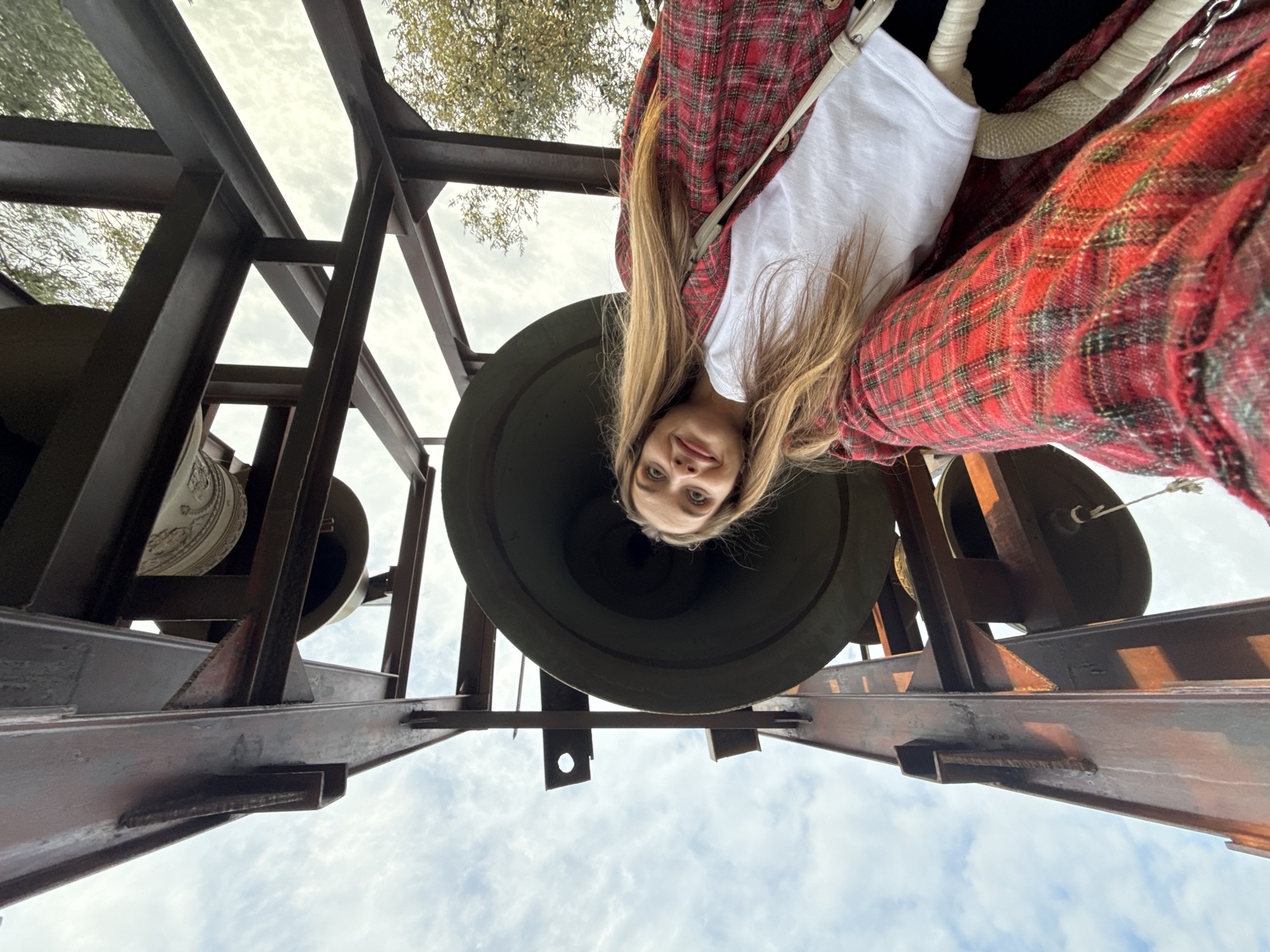 A low-angle, upside-down shot reveals a young woman positioned directly beneath a large, dark-colored bell within a metal framework. She is wearing a white shirt and a red and black plaid jacket. The metal structure supporting the bell has a brown, possibly rusted, appearance. Additional bells are visible behind and to the sides of the primary one. The sky, partially obscured by the framework and trees, is visible in the background. The location is Đuraševići, Montenegro.