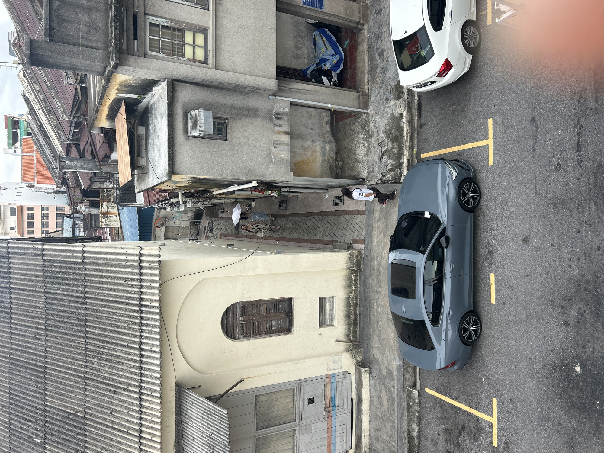 This is an elevated, eye-level, medium shot captured in George Town, Malaysia, depicting a narrow alleyway flanked by weathered, multi-story buildings. On the left, a building with a corrugated metal roof and a cream-colored facade features a large, arched wooden window with visible louvers and a smaller rectangular vent below. To the right of this building, a narrow paved alleyway leads into the distance.
In the alleyway, two women are walking away from the camera, one holding a white umbrella. Further down, a young woman in a white t-shirt and dark pants is walking towards the camera. The buildings on the right side of the alley are constructed of concrete and exhibit signs of age and wear, with stained walls and aging windows. One ground-level opening on the right shows a motorcycle covered with a blue and white tarp.
In the foreground, two cars are parked on an asphalt surface marked with yellow parking lines. A dark gray sedan, possibly a Volvo, is parked on the left, and a white sedan is parked on the right. The overall scene conveys a sense of an older, urban environment with a mix of residential and commercial structures.
