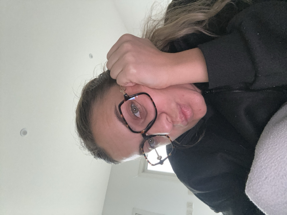 An indoor, close-up image taken in Veauchette, France, displays a female subject from the chest up. She wears a dark-colored, zippered top and large, square, tortoiseshell-patterned eyeglasses with gold accents at the temples. Her light brown hair is partially visible, and her eyes are directed forward. She rests the right side of her face on her right hand, with her fingers visible near her temple, while her lips are pursed in a puckered expression. A light-colored fabric is visible in the lower left foreground. The background consists of a plain, light-colored wall with a circular ceiling fixture and rectangular wall fixtures visible to the left.