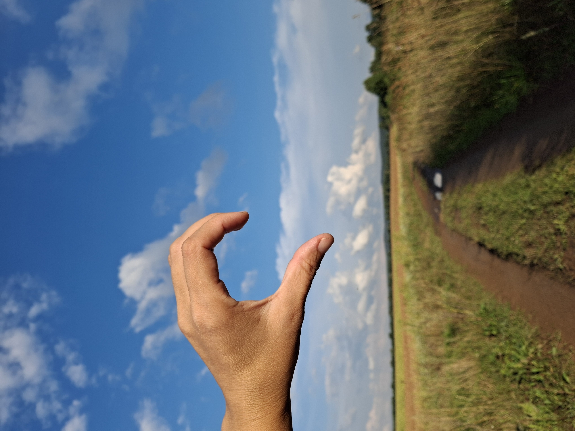 An outdoor, eye-level, medium shot features a human hand forming a heart shape against a bright blue sky with scattered white clouds. The hand is positioned in the foreground, on the left side of the frame, with the fingers curled inward and the thumb extended. The background shows a dirt path winding through a field of tall, dry grass and some greenery. In the distance, a blurred line of trees and hills can be seen under a partly cloudy sky. The lighting suggests it is daytime, with the sun casting soft shadows. The image does not provide any identifiable landmarks or details that would specify its location within Plishkino, Russia.