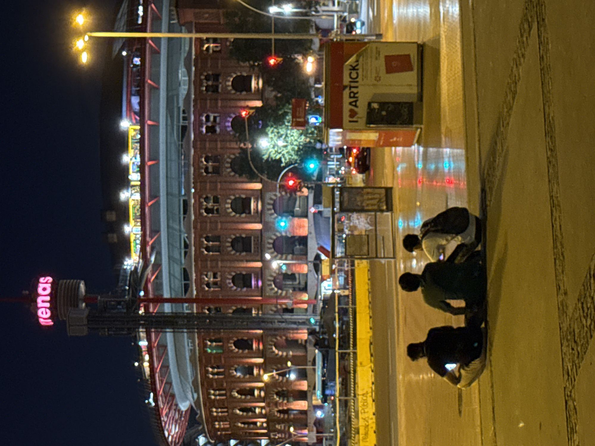 The image depicts a nighttime street scene in Barcelona, Spain, focused on the Arenas de Barcelona, a former bullring converted into a shopping center, with the iconic "arenas" sign prominently lit in pink. In the foreground, three individuals are seated on the ground, facing away from the camera, appearing to be looking at their phones. The ground is wet, reflecting the colorful lights of the city, suggesting recent rain. Streetlights and traffic signals add to the urban ambiance. The architecture of the Arenas is visible in the background, characterized by its circular shape and brick facade, illuminated with warm-toned lights. The overall impression is a calm, urban setting at night, with people pausing in their activities amidst the city's glow.