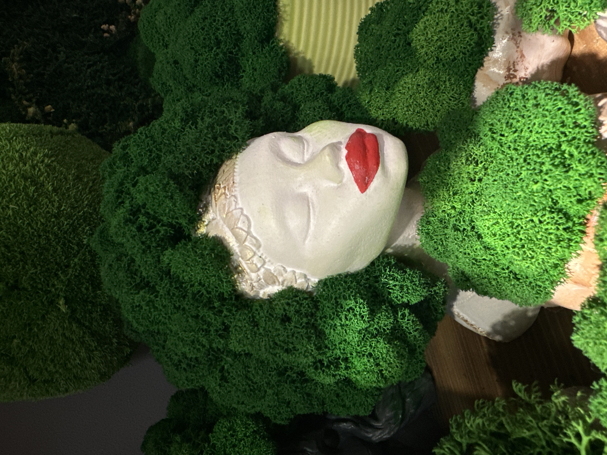The image depicts a white sculptural bust of a woman, facing right with closed eyes and bright red lips. Her head is adorned with bright green, voluminous moss simulating hair, featuring a textured golden band at the hairline. The bust is surrounded by additional green moss formations, some lighter and some darker, consistent with decorative elements within an interior setting in Rostov-on-Don, Russia. A dark, partially obscured sculptural object is visible in the background to the left. To the right of the bust, a light green, horizontally ribbed surface is present. The entire arrangement rests on a dark, wood-textured surface, with smaller, moss-covered beige and light-colored rocky elements visible in the foreground. The scene is illuminated, creating distinct shadows.