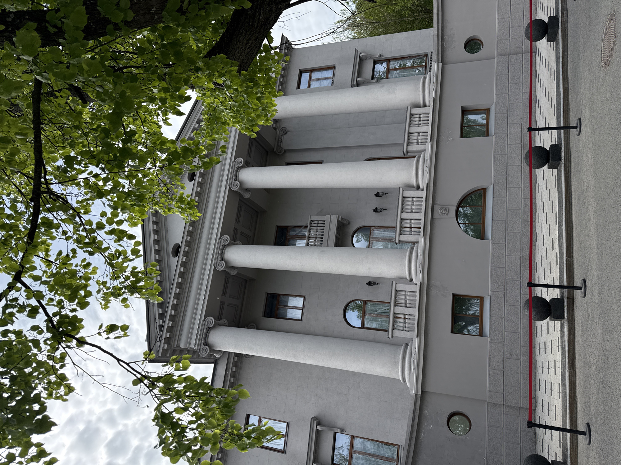 The image shows a three-story building with classical architectural features, likely located in Nizhny Novgorod, Russia, based on architectural style. The building is light gray with prominent white columns on the facade. Some of the windows are rectangular, while others are arched. The building has decorative details such as sculpted elements and railings on small balconies. A tree with green leaves partially obscures the view of the building on the left side. A walkway with gray paving stones and black bollards connected by a red rope runs along the building's right side. The photo is taken from a low angle, looking up at the building, with the sky visible through the branches of the tree.