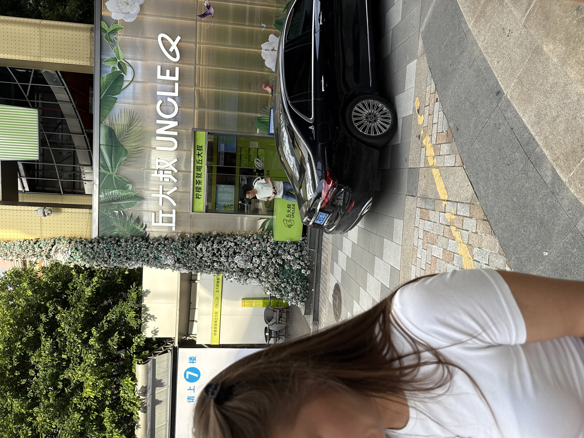 In Haizhu District, China, a woman with long brown hair tied back with a dark hair clip stands in the foreground, partially obscuring a street scene. She wears a white t-shirt. Behind her, a black Mercedes-Benz sedan is parked on a sidewalk with a distinctive patterned stone paving. Adjacent to the car is a modern building with a translucent facade displaying large green foliage graphics. Above the entrance to a business, the words "UNCLE Q" are prominently displayed in large white lettering, with smaller Chinese characters above and below. To the left of the "UNCLE Q" signage, a tall, narrow artificial Christmas tree-like structure is decorated with numerous small white and teal-colored blossoms and silver flowers. In the background, a person wearing glasses and a light-colored t-shirt and blue jeans stands near the entrance of "UNCLE Q," holding a mobile phone. A surveillance camera is visible on a yellow-clad structure to the upper left of the scene. The street is paved with square grey and beige tiles and has a yellow painted marking on it.