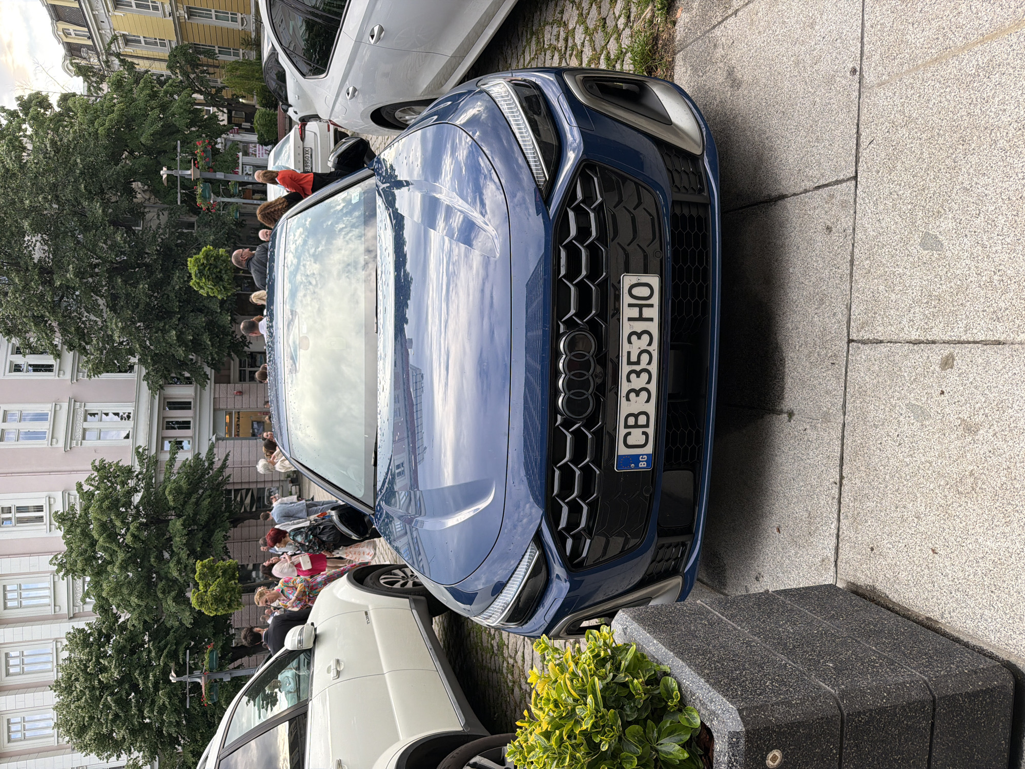 A blue Audi passenger car with license plate CB 3353 HO is parked at a curb on a city street. The photograph is shot at street level. In the background, people are walking along the sidewalk in front of buildings. The country code on the license plate indicates the location is Bulgaria. Burgas is assumed based on metadata. There is a white car parked next to the Audi. In the foreground, there is a planter box. The street appears to be paved with stone.