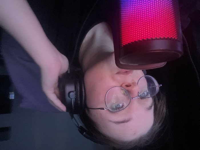 A young adult female is depicted in a close-up, indoors, under low light conditions. She has light skin, dark hair, and wears round, wire-frame glasses with light-colored lenses. Her eyes are directed upwards and to the viewer's left. She wears black over-ear headphones, with her left hand holding the left earcup against her ear. Her mouth is slightly open. In the lower-left foreground, a cylindrical microphone with a mesh grille displays a gradient of red, pink, and purple illumination. The individual is wearing a dark, presumably black, top. The background is dark and indistinct, with a dark blue/purple fabric visible behind her right shoulder. The specific location within Pushchino, Russia, cannot be determined from the visual information provided.