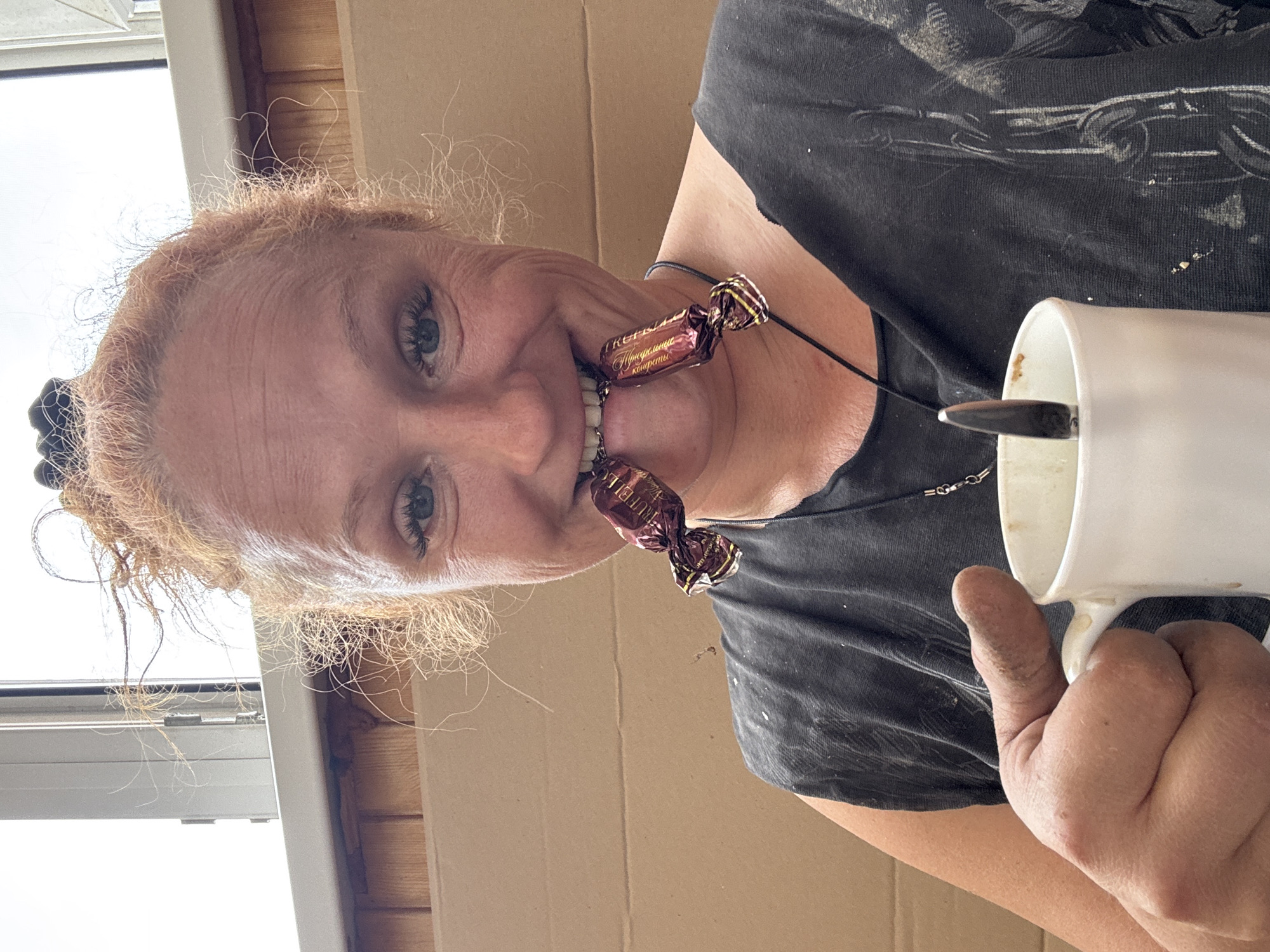 A middle-aged Caucasian woman with red hair is seen from a low, tilted angle. She is holding a white mug, likely containing coffee, with a spoon visible inside. She has two wrapped candies ("Trufel") in her mouth as if she is wearing them as a necklace. She is smiling. The background includes a wall with a window. This image was taken in Chaikovsky, Russia. The woman is wearing a black t-shirt. Her hand holding the mug appears to be dirty, possibly from working or another activity.