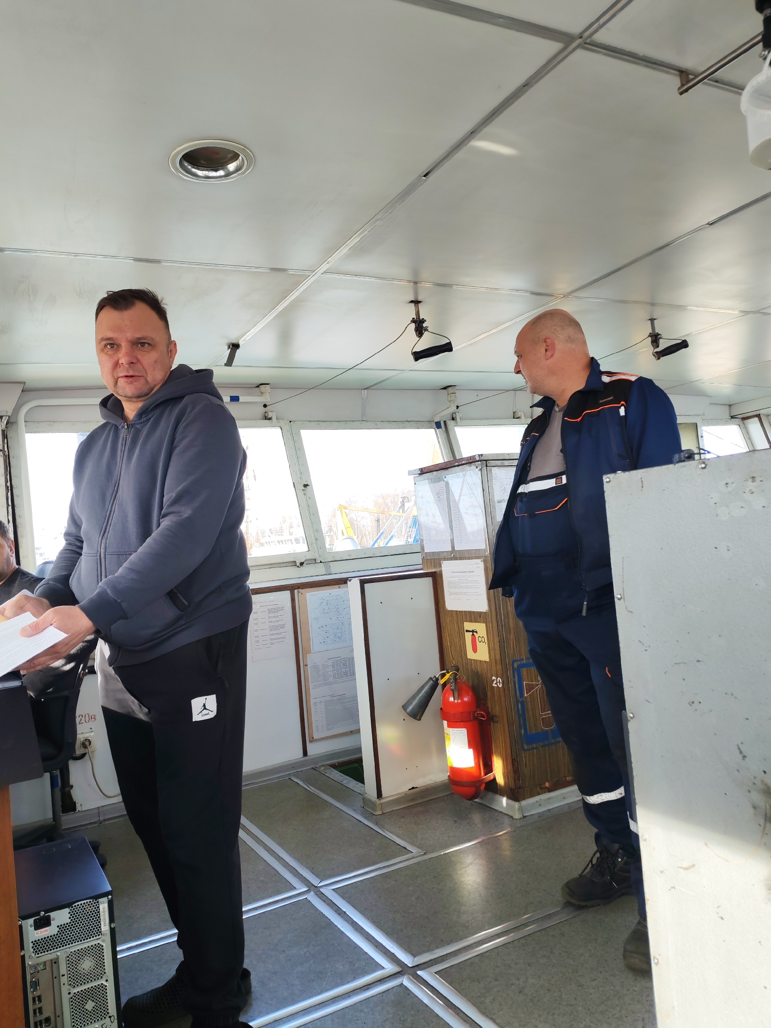 The image shows the interior of what appears to be the wheelhouse of a vessel, possibly a riverboat, with two men inside. The location is suggested as Rybinsk, Russia.
One man, wearing a dark blue hooded sweatshirt and dark track pants with a white logo near the left thigh, is standing on the left. He's holding a stack of papers in his hands.
The second man is on the right, wearing a dark blue work suit with orange accents. He's looking to the left.
The wheelhouse has large windows providing a view of the outside. Wall mounted information boards are visible in the background.
A red fire extinguisher is mounted on a wooden cabinet near the center of the frame. The floor is made of gray tiles with metal strips delineating the tile boundaries. The ceiling is white and features an overhead light fixture. A computer tower is visible at the base of the man in the hoodie.
