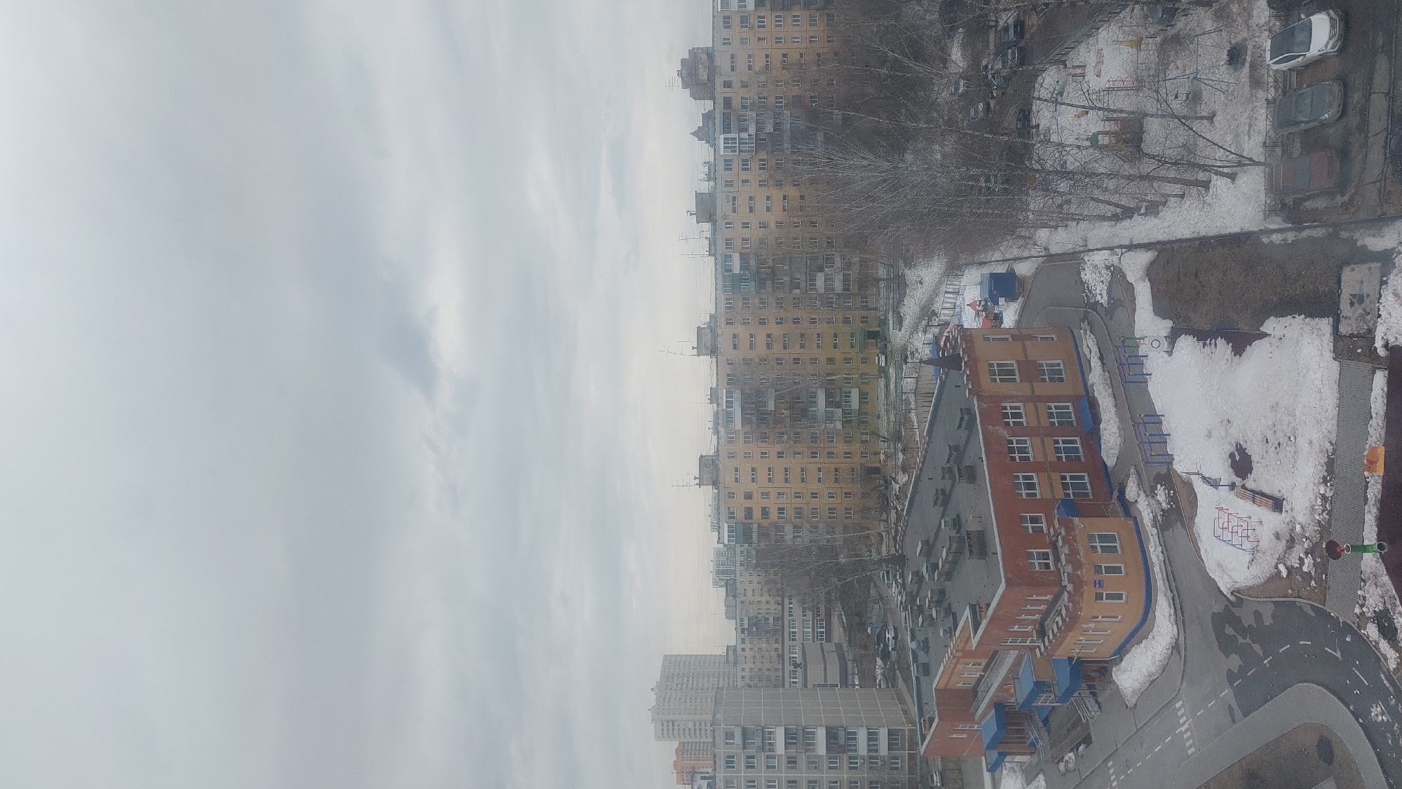 This is an aerial view of a residential area in Novosibirsk, Russia, likely taken from a high-rise building. The scene depicts a cloudy sky above a cityscape dominated by apartment buildings of varying heights and architectural styles. Snow is visible on the ground in patches, particularly around a playground area and on unpaved surfaces. A newly built school or kindergarten building can be seen next to a playground with a road simulation area for teaching the rules of the road. Cars are parked in designated areas. The environment suggests an urban setting experiencing the transition between winter and spring. No people are visible in the frame. The overall atmosphere is cold and overcast.