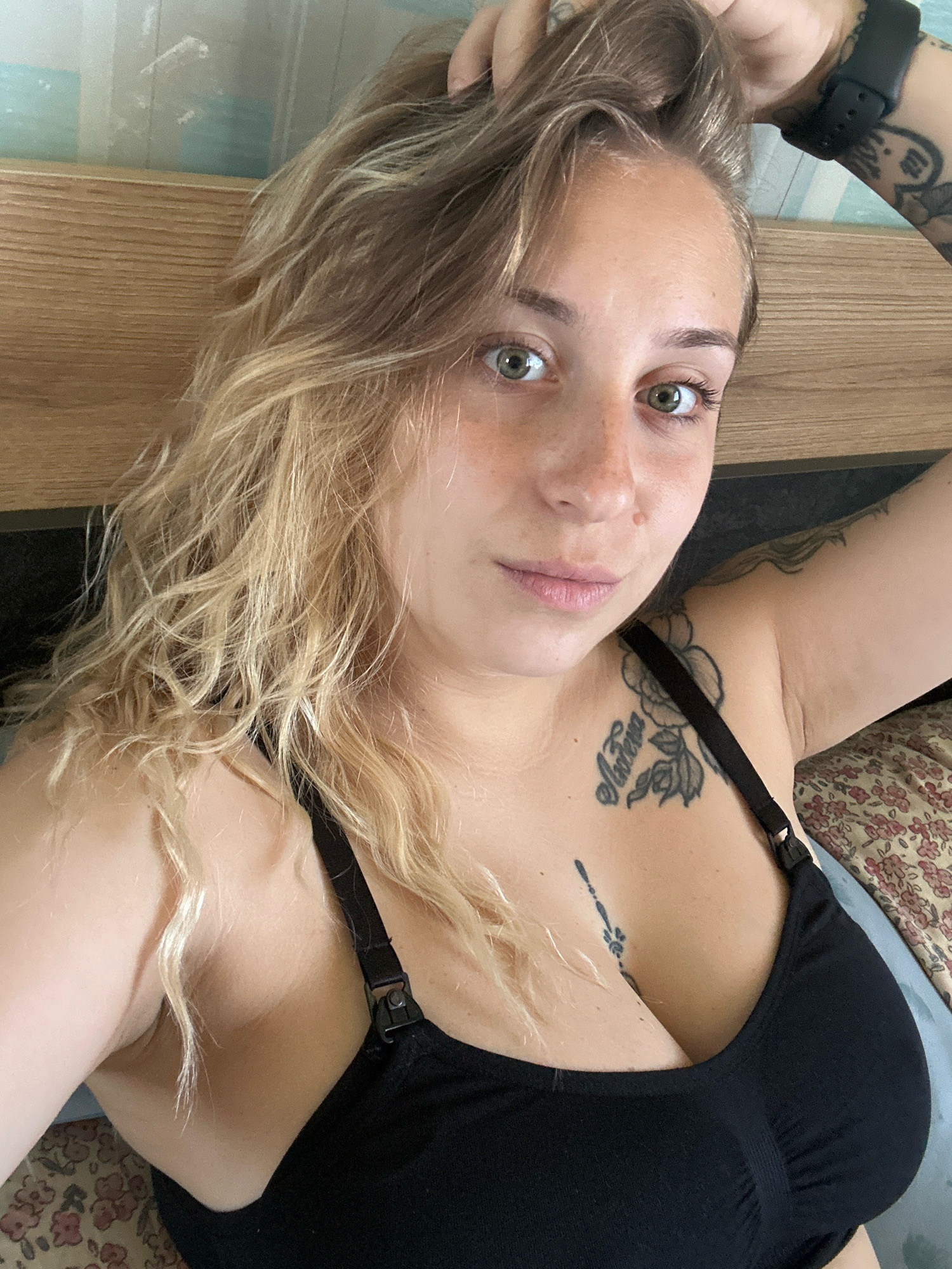 A woman with blonde hair and green eyes, wearing a black bra, is lying down. She has tattoos on her arm and chest. The background appears to be a bedroom with a wooden headboard and a floral patterned bedspread. There is no visible indication of the image being taken in the city of Plovdiv, Bulgaria.