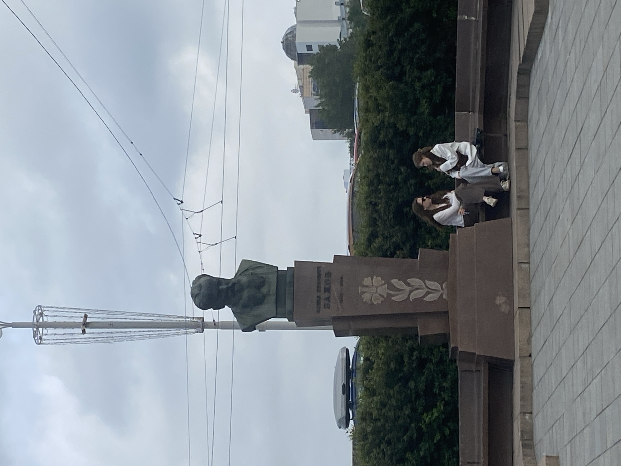 The image depicts a monument dedicated to Pavel Bazhov, a Soviet and Russian writer, located in Yekaterinburg, Russia. Two young women are seated on a bench in front of the monument, engaged in conversation. The monument itself features a bust of Bazhov atop a stone pedestal, which is adorned with an engraved floral motif and the writer's name and birth and death dates. Behind the monument, a dense hedge forms a backdrop, with a car equipped with a roof box visible to the left. In the distance, several buildings, including one with a domed structure, are silhouetted against a cloudy sky. Overhead, power lines and a street light pole are also visible. The ground in the foreground is paved with square tiles. The overall scene suggests a public urban space, likely a park or square, on an overcast day.