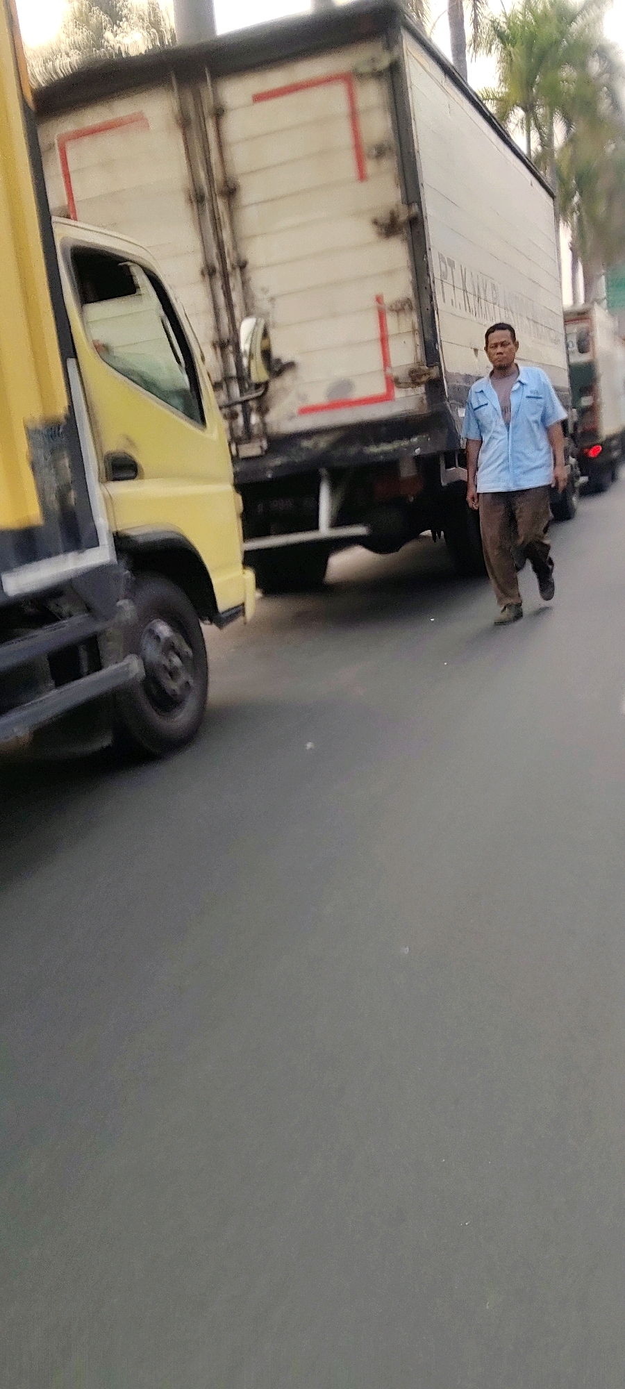 In Mekarmukti, Indonesia, a man walks on a street next to a line of parked trucks. The man, wearing a blue collared shirt and brown pants, is walking away from the camera, his right leg forward. He appears to be walking between two trucks, one yellow and one white with red markings. The white truck has text printed on its side that reads "PT. KAKAK PER". The yellow truck is partially visible on the left side of the frame. The ground is paved and appears to be asphalt. Trees and palm trees are visible in the background, suggesting an outdoor urban environment.