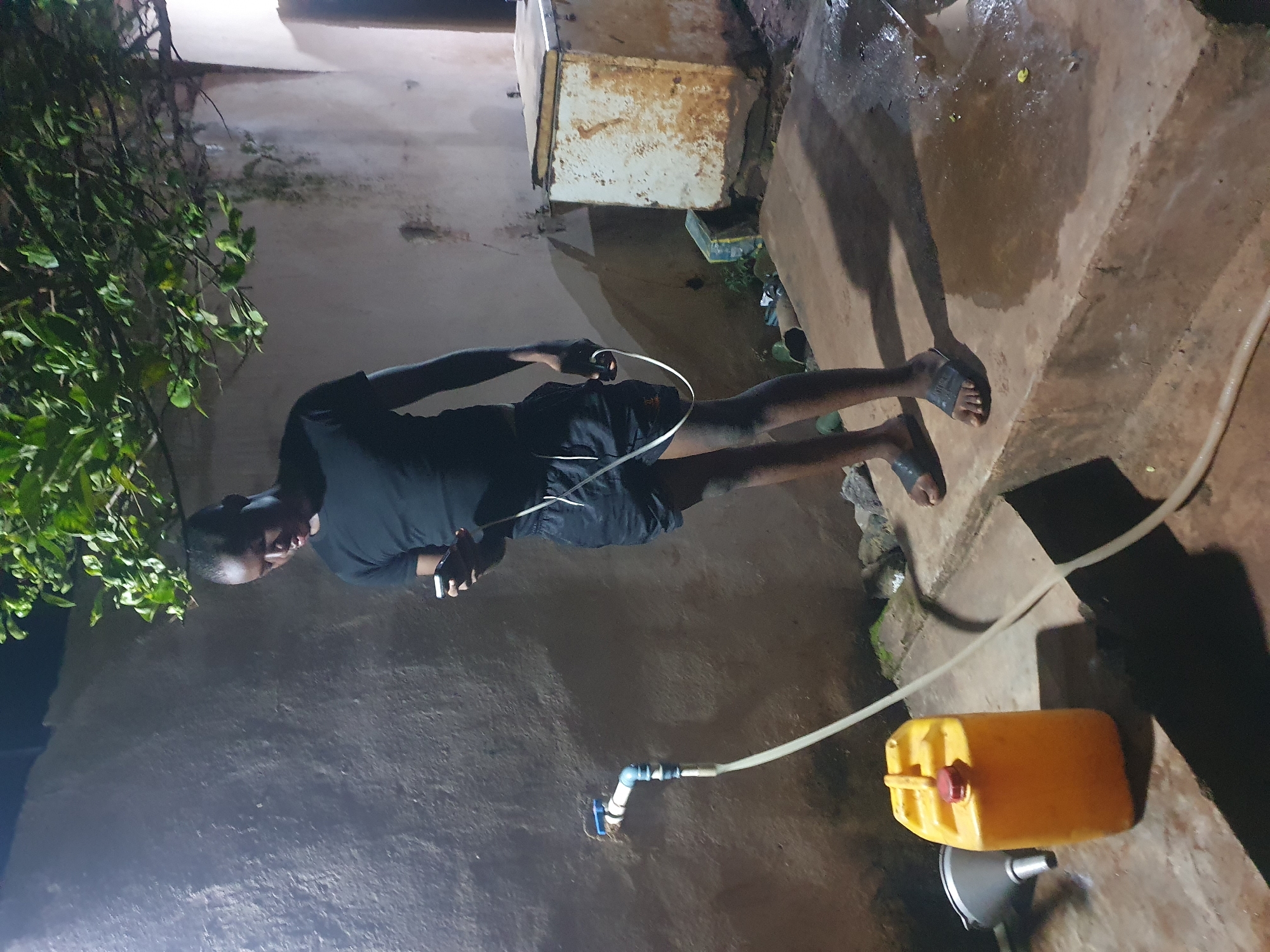 The image shows a man standing outdoors at night in what appears to be a residential area in Idah, Nigeria. The man is wearing a dark t-shirt, shorts, and sandals, and is looking at a mobile phone in his hands. A white cord is visible, possibly from the phone or earphones. Next to him, a yellow plastic container is connected to a water faucet via a hose and some blue and silver fittings. A funnel sits nearby, likely to help in filling the container. The area is paved with what appears to be weathered concrete. A rusted metal cabinet and foliage are visible in the background. The area appears to be dimly lit.