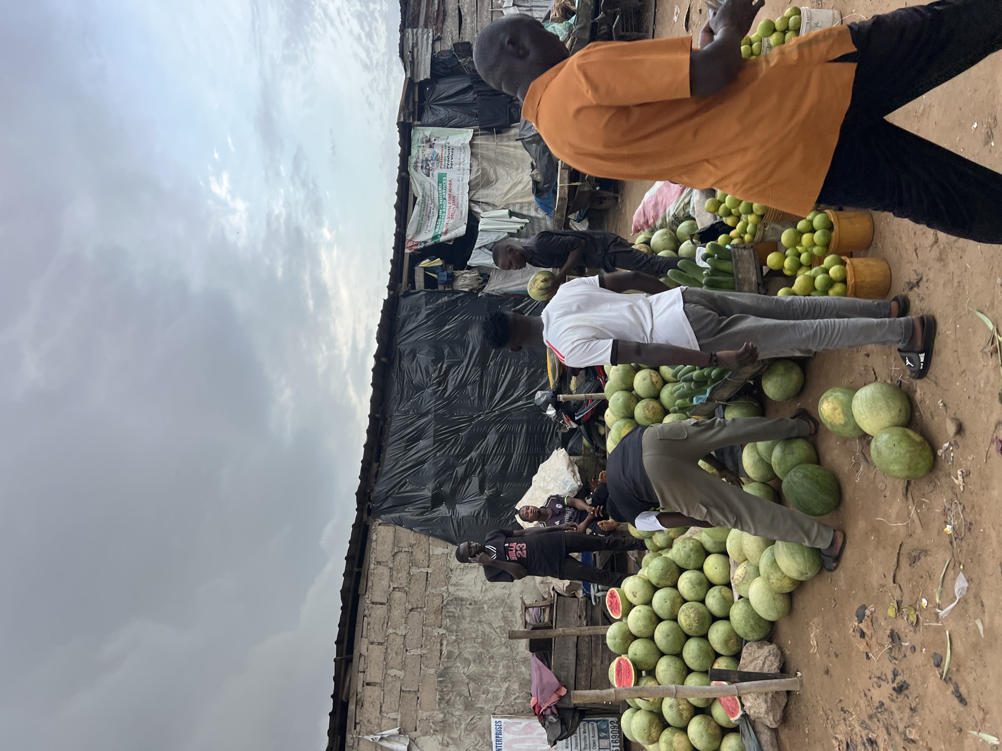 In Asaba, Nigeria, a bustling street market unfolds under an overcast sky. Piles of ripe watermelons dominate the foreground, their green rinds contrasting with the earthy brown dirt. Several men are engaged in the market's commerce. One man, wearing a black t-shirt with "BULLS 23" emblazoned on it, is speaking on a mobile phone, standing amidst the produce. Nearby, another man in a white t-shirt is interacting with a display of watermelons, and a third man in an orange t-shirt walks away from the camera. Behind them, stalls are constructed from cinder blocks and covered with black tarpaulin, suggesting a semi-permanent marketplace. Signs advertising various enterprises and government initiatives, some in English and some in local languages, are visible on the structures. The scene captures a moment of everyday life in an open-air market, with the sale of fresh produce being the central activity.