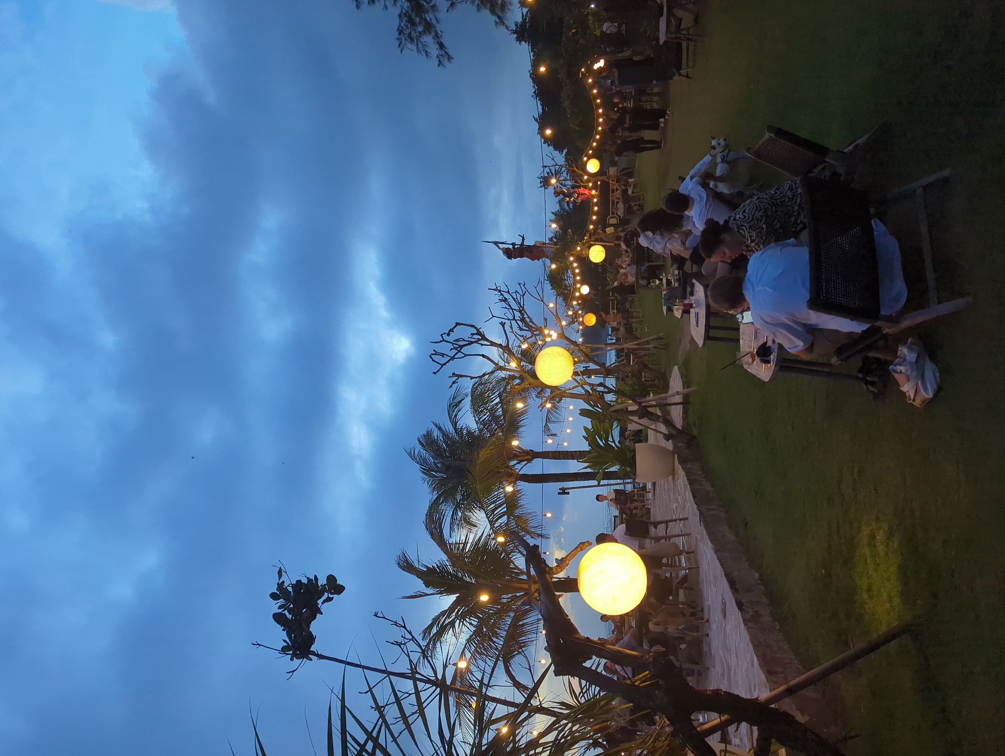 At dusk in Mengwi, Indonesia, an outdoor dining area is illuminated by spherical string lights, creating a warm ambiance. Patrons are seated at tables, some engaged in conversation. Palm trees add to the tropical feel, while a statue is visible in the background. The sky is cloudy, with shades of blue and gray.