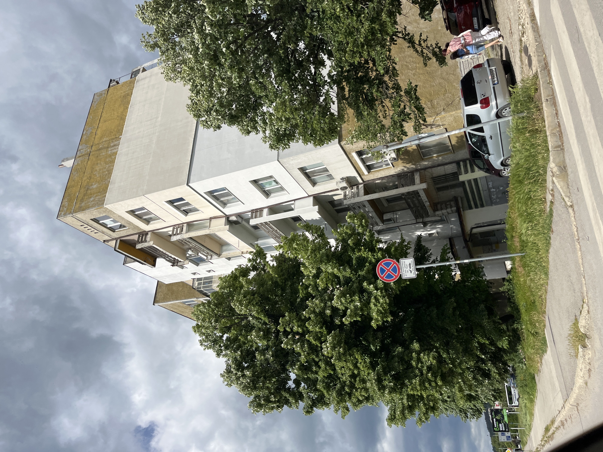 The image shows the exterior of a multi-story building in Varna, Bulgaria. The building has a light yellow and white facade with several windows and balconies. A large tree partially obscures the lower portion of the building, and a no-parking sign is visible in front of the tree. A street runs alongside the building, with a white car and several people visible. The sky is cloudy and overcast.