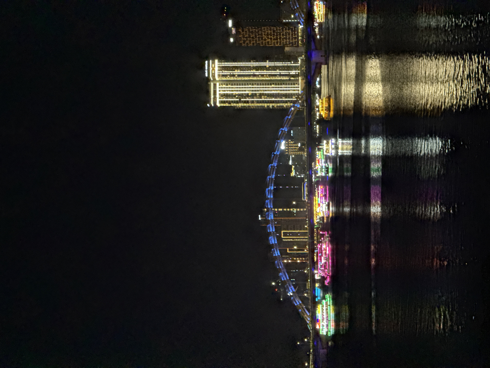 The image depicts the Han River at night in Da Nang, Vietnam, showcasing the Dragon Bridge and illuminated buildings. Several tour boats, adorned with vibrant neon lights in green, pink, and yellow, traverse the dark water, their reflections shimmering on the surface. In the background, two tall buildings stand out, one with a prominent "HELLIX VUALI" sign, both illuminated with vertical lines of light. The Dragon Bridge itself is a prominent feature, with its arched structure outlined in blue lights. The overall scene is a nighttime cityscape, emphasizing the urban infrastructure and water-based tourism in Da Nang.