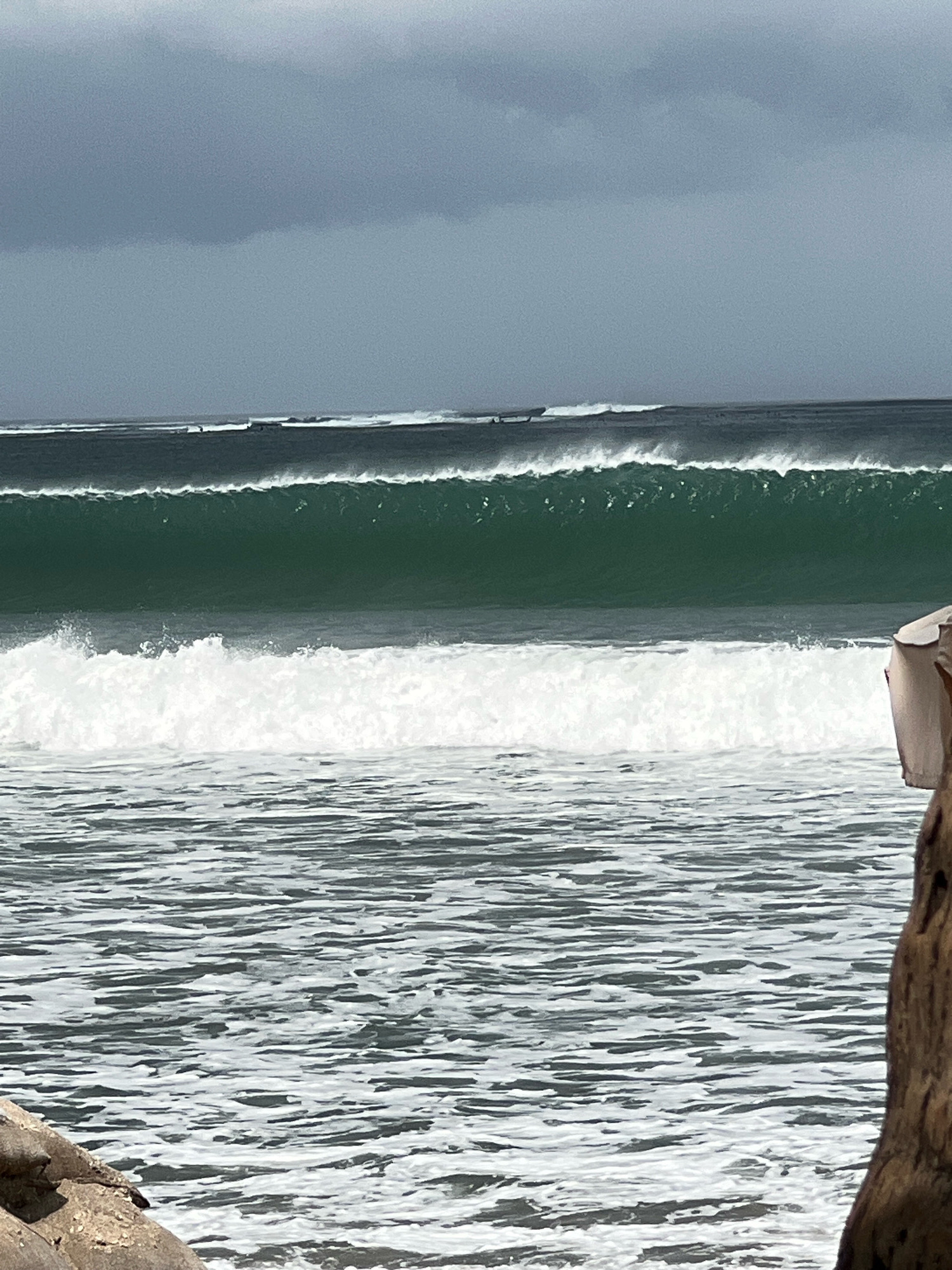 The image shows a cloudy, overcast day at Kuta Beach, Indonesia, with large waves rolling in. The water is a deep green, and the waves are breaking with white foam. In the distance, several surfers can be seen on the water. The foreground of the image shows a portion of a large piece of driftwood, partially framing the scene.