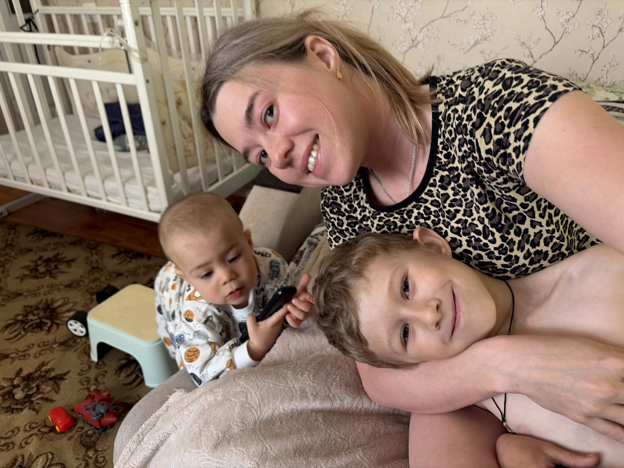 The image shows a woman with blonde hair and a leopard-print top smiling at the camera. She is embracing a young boy with light brown hair. To their left is a baby wearing a patterned pajama. The baby is holding a black remote control. A small, light green stool with a tan top is next to the baby. A red toy car and a red and grey toy truck are on the carpet below the stool. Behind them is a white crib. The backdrop features patterned wallpaper. The location is implied as Chaikovsky, Russia.