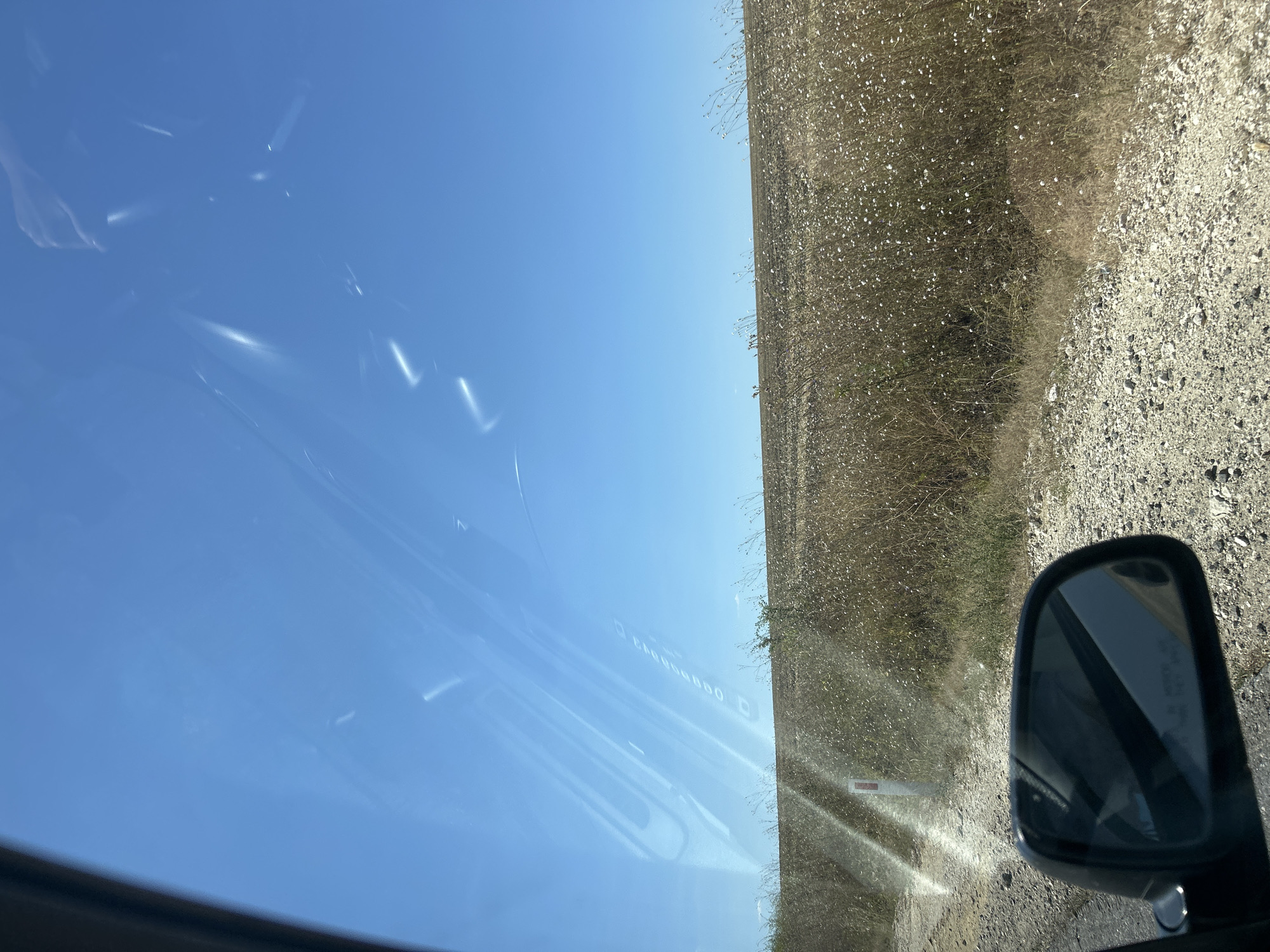 The image captures a view from inside a vehicle, looking out through the windshield. The sky is a clear, bright blue, with some subtle reflections and streaks visible on the glass, suggesting it might be a sunny day. A portion of the car's side mirror is visible in the lower left corner of the frame. Beyond the glass, a dry, grassy field stretches towards the horizon. The terrain appears somewhat rugged, with a gravel road leading into the scene from the left. The vegetation in the field is predominantly dry grass and small white wildflowers. In the background, the land appears cultivated or fallow, with a gentle slope. A white post with a red stripe, likely a road marker, is visible on the left side of the gravel path. The overall impression is of a rural landscape under a vast, open sky. The scene is consistent with a location in the vicinity of Comrat, Moldova, given the agricultural nature of the landscape and the presence of typical roadside markers.