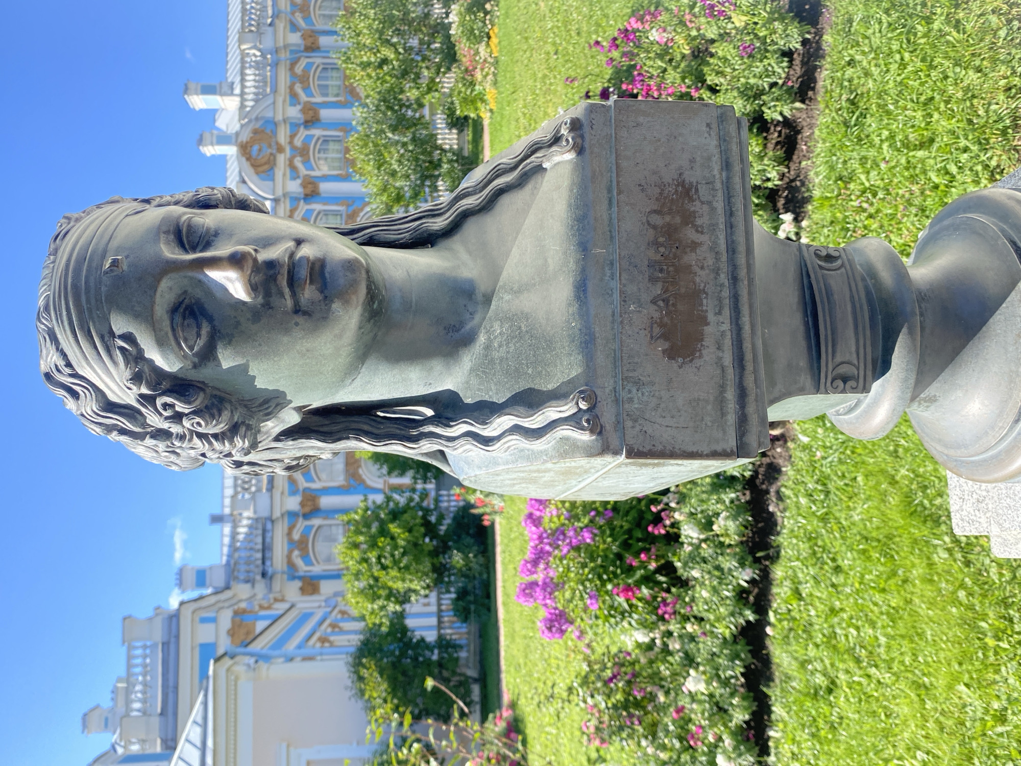 The image shows a bronze bust of a woman with long, wavy hair, wearing a headband. The bust is positioned in the foreground, on a pedestal, with a garden and a palace in the background. The palace, painted in white and blue with gold accents, is characteristic of Baroque architecture and is likely the Catherine Palace in Pushkin, Russia. The garden features lush green grass and blooming pink and white flowers. The sky is a clear, bright blue. The bust appears to be an outdoor sculpture in a park or garden setting. The inscription on the base of the bust is partially visible but unreadable.
