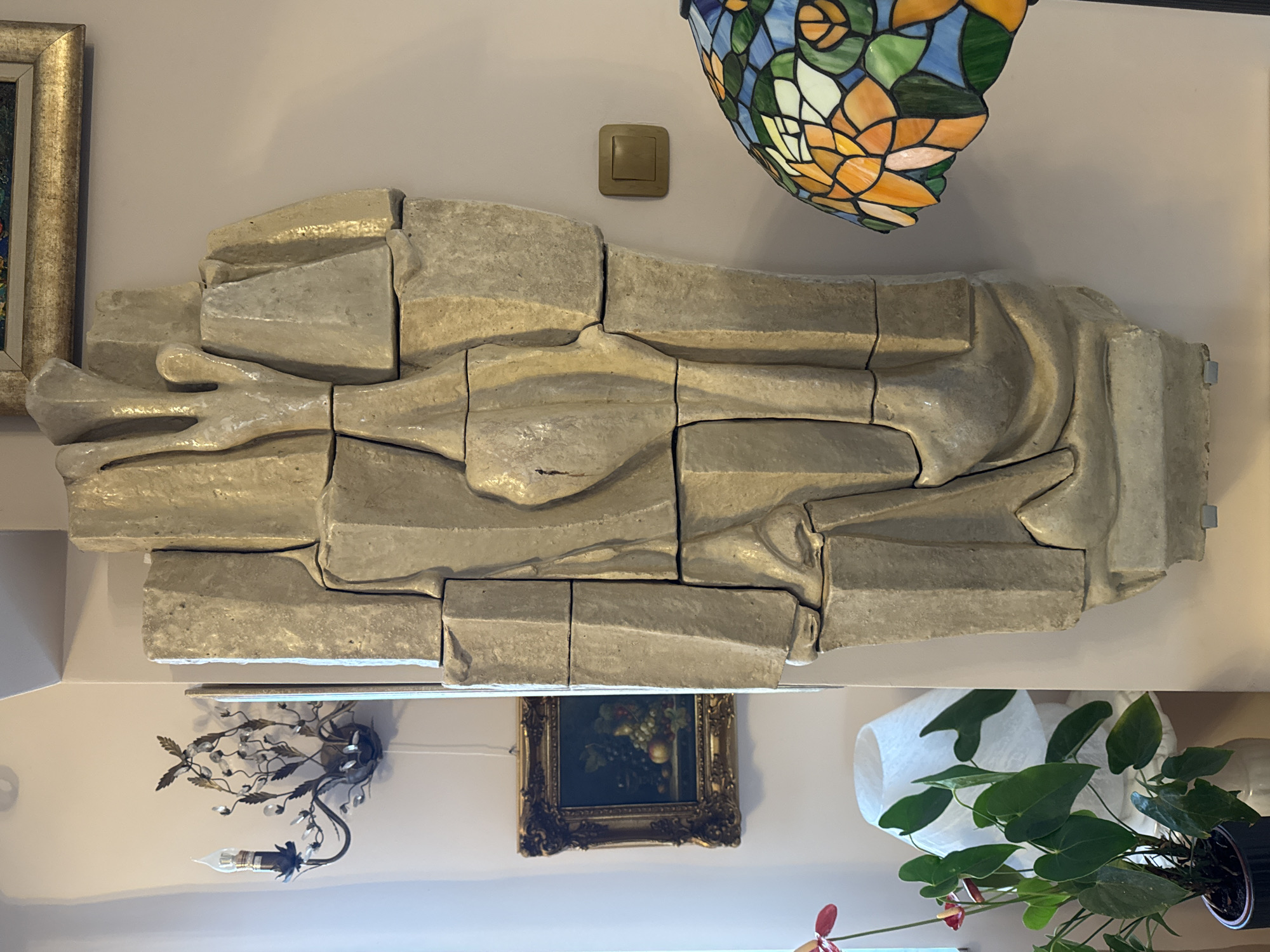A horizontally oriented medium shot shows a light gray sculpted stone art piece, possibly depicting a reclining figure, mounted on a beige-colored wall. The sculpture is composed of multiple stone blocks that are fitted together. Above the sculpture, a square electrical outlet plate and a multi-colored Tiffany-style lamp fixture are visible. Below the sculpture, a vintage-style wall sconce, a framed painting of fruit, a lamp with a white shade, and a potted plant with large green leaves are placed. The overall setting suggests an interior space, possibly a residential home. There is no verifiable information in the image to confirm that the location is in Plovdiv, Bulgaria.