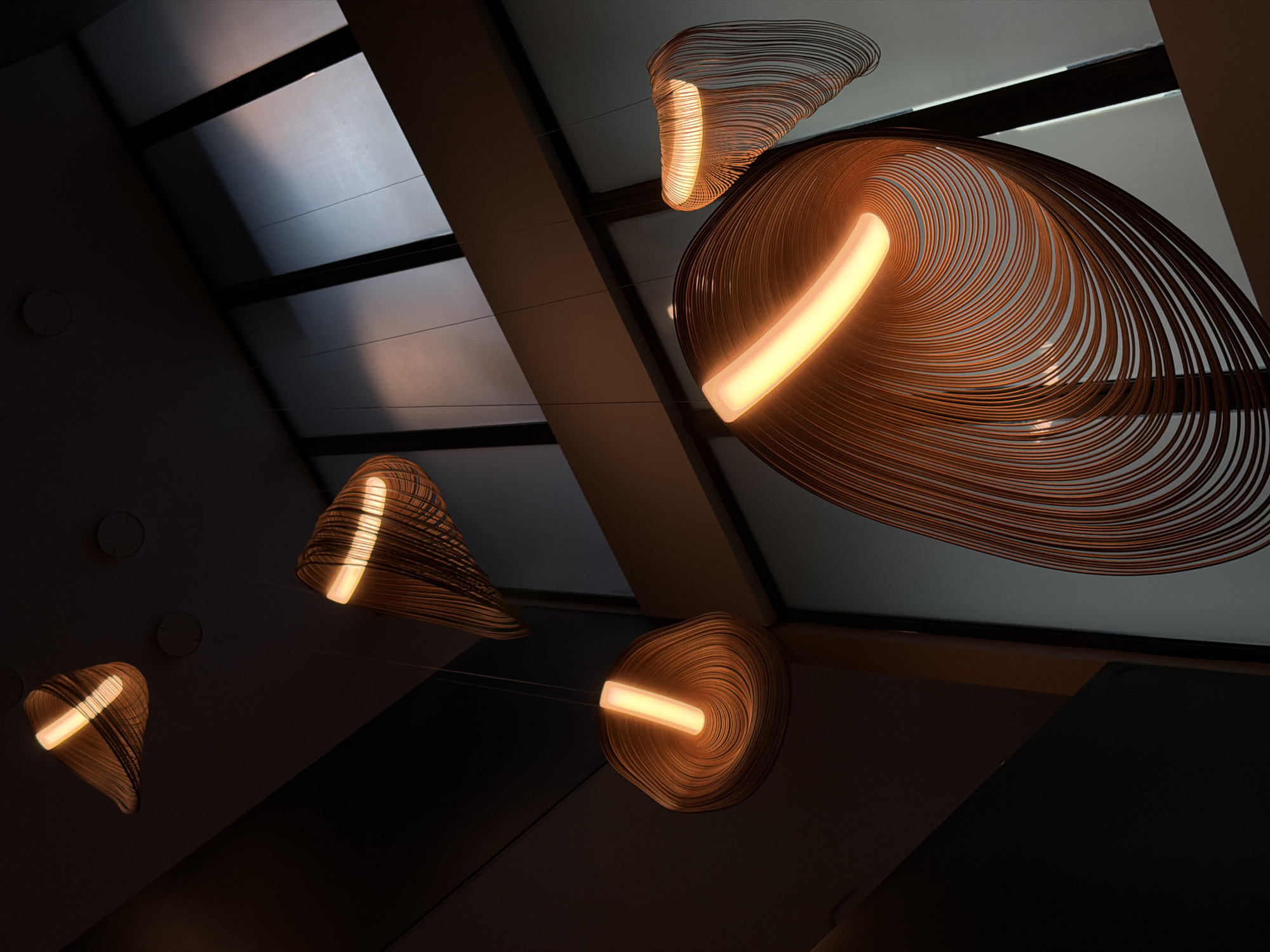 The image depicts several modern pendant lights suspended from a sloped ceiling with visible skylights. The pendant lights feature a unique design of multiple thin, curved strips arranged in an organic, almost fluid shape. Each light has a central illuminated tube, casting a warm, inviting glow. The ceiling has a geometric pattern of panels and supporting beams. The overall ambiance is warm and contemporary. The exact location in Kuta, Indonesia, is unknown from the provided image.