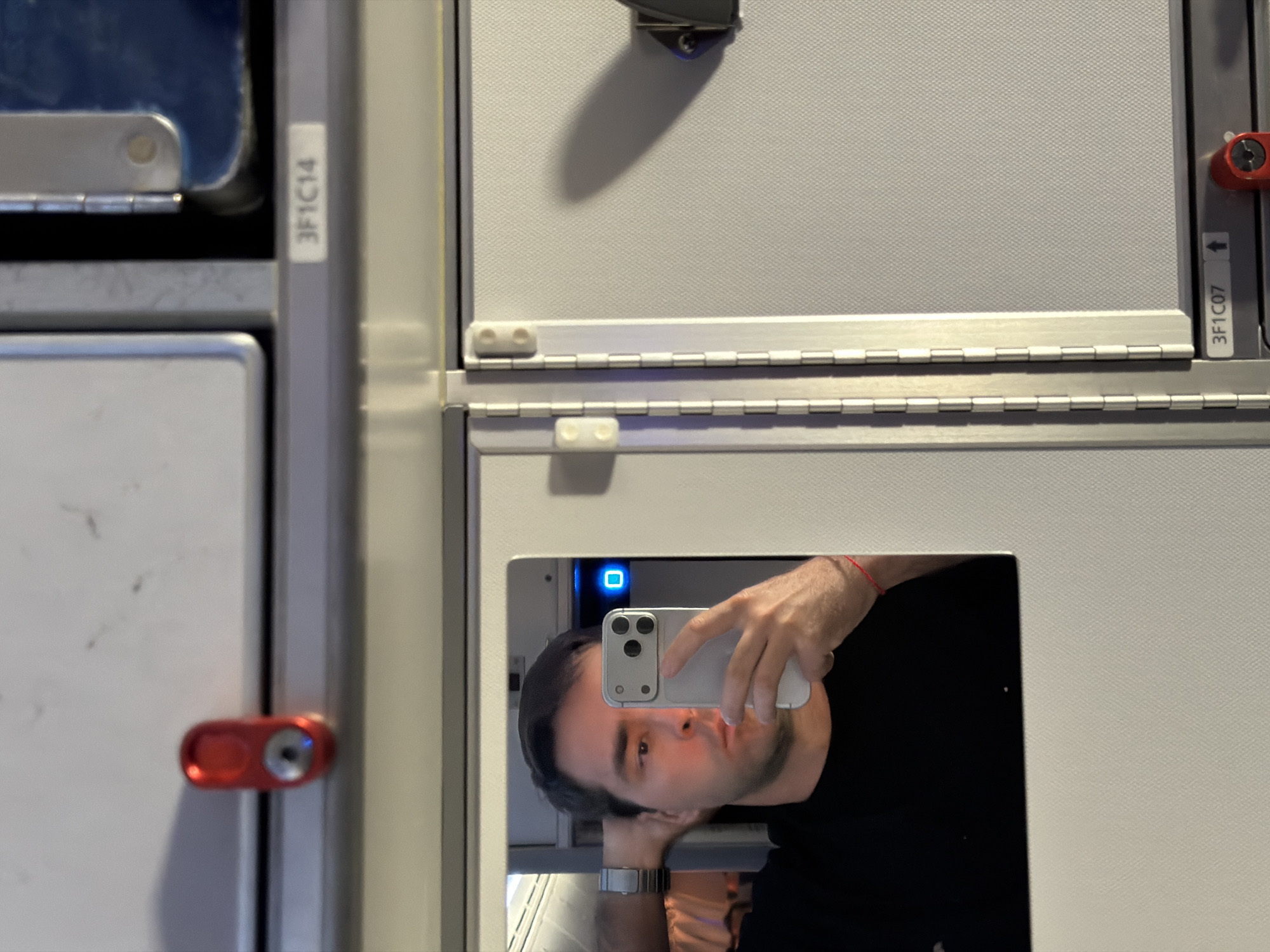 A man is taking a selfie in a mirrored surface. The mirror appears to be part of a structure in an airplane lavatory. The man is holding a white smartphone, with the camera lenses visible. He is wearing a black shirt and a wristwatch on his left wrist. The mirrored surface reflects details of the lavatory, including the white walls and fixtures. The numbers "3F1C14" and "3F1C07" are labeled on the walls. It is not possible to determine the location in the city of Tzafria, Israel.