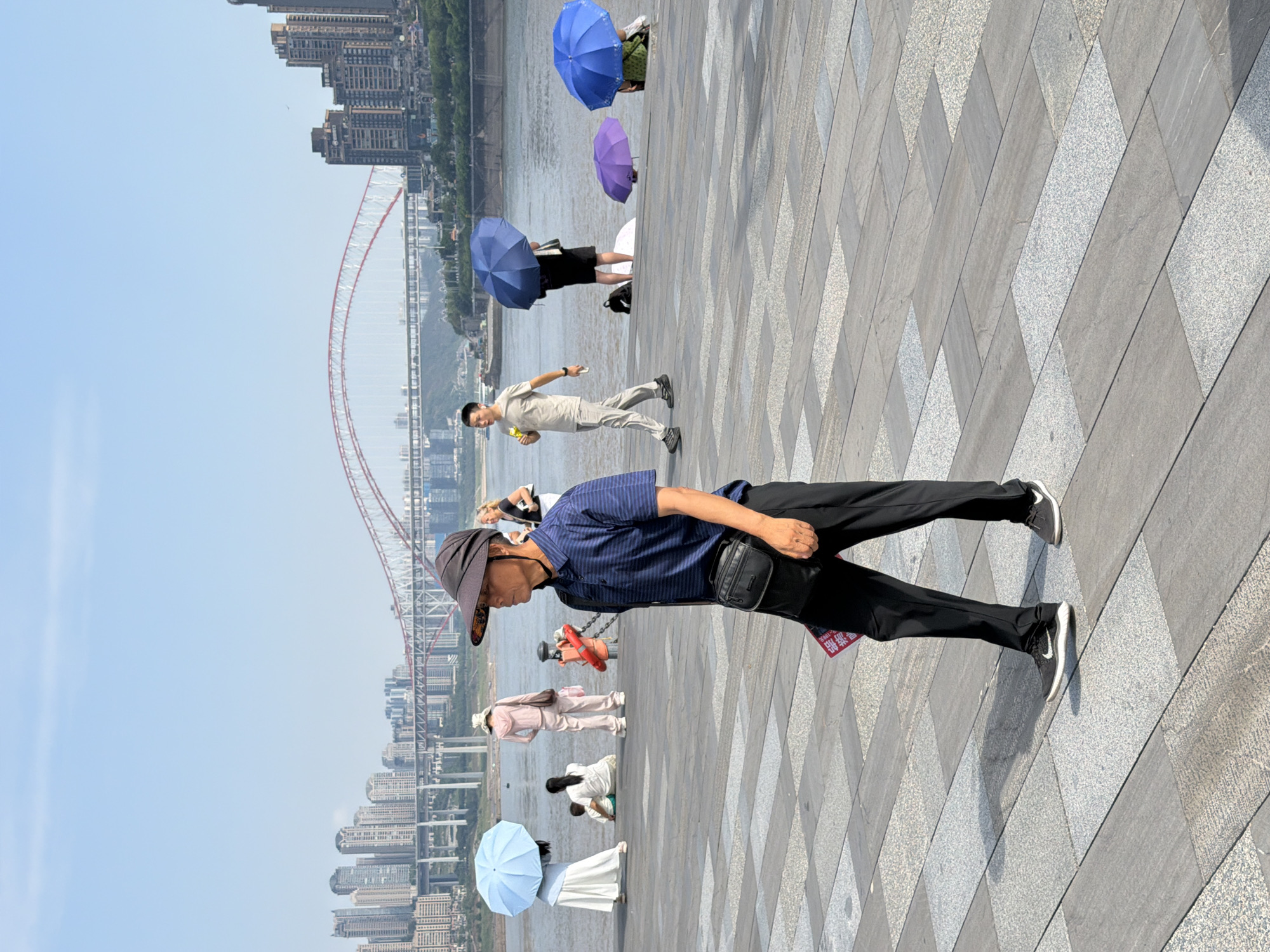 This image captures a sunny day along the waterfront in Nan'an District, Chongqing, China. In the foreground, a man wearing a wide-brimmed hat, a blue striped polo shirt, black pants, and a cross-body bag walks on a paved walkway. He is holding a red pamphlet in his left hand. Behind him, several other people are visible near the water. A woman with a light blue umbrella stands on the walkway, and two children are seated nearby. Further back, a person in a pink outfit walks away from the camera, and a man in a light-colored shirt and pants walks toward the river, holding a yellow object. Several colorful umbrellas, blue and purple, are open and held by people standing by the water's edge. In the background, the Yangtze River flows, with a large red-arched bridge spanning across it. The cityscape of Nan'an District is visible on the far bank, with numerous tall buildings and structures lining the shore. The sky is clear and blue.