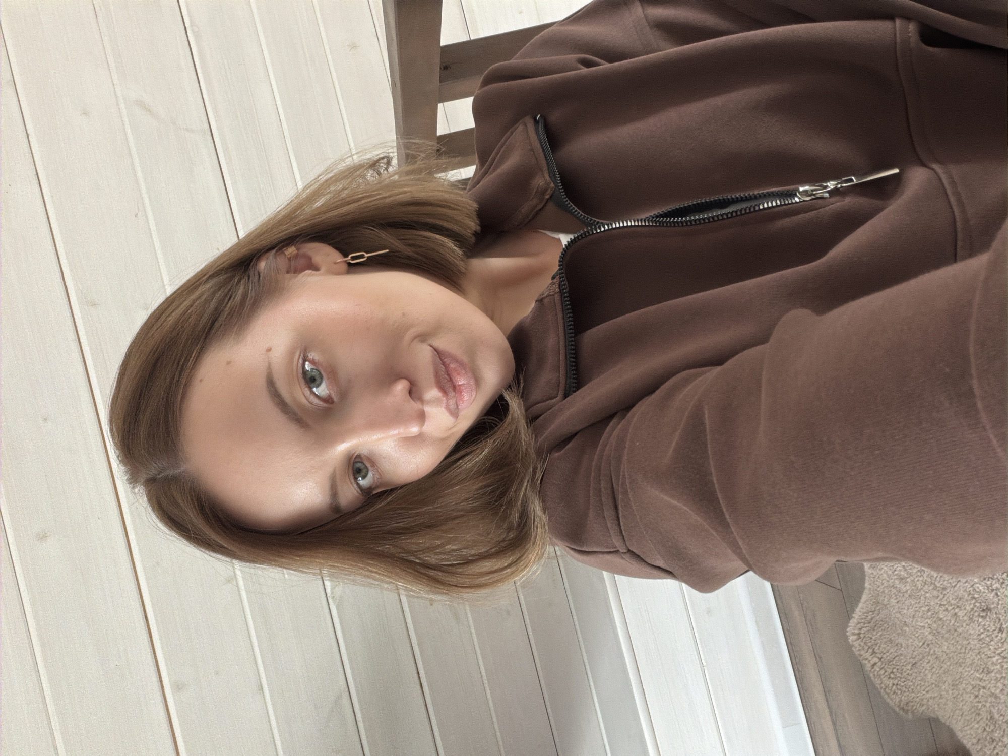 A young woman is depicted in a close-up, selfie-style portrait, looking directly at the camera with a calm, soft gaze. The image is tilted, suggesting a casual, spontaneous shot. She has short to medium-length light brown hair, parted on her left, and light-colored eyes that appear green or hazel. Her lips are slightly parted, and she has minimal, natural-looking makeup.
She is wearing a brown, full-zip hooded sweatshirt or jacket, partially unzipped to reveal a glimpse of a light-colored garment underneath. A small, elegant, gold-toned earring with a chain-link design is visible in her right ear.
The background features light-colored, vertically oriented wooden wall paneling, suggesting an indoor setting. A dark wooden chair or piece of furniture is visible behind her head on the right side of the frame. In the lower right portion of the image, a section of a textured, light beige carpet is present. The lighting is soft and even, indicating a daytime setting. The scene appears to be a casual, domestic interior, possibly in Бунятино, Russia, based on the provided context. No visible text is present in the image.