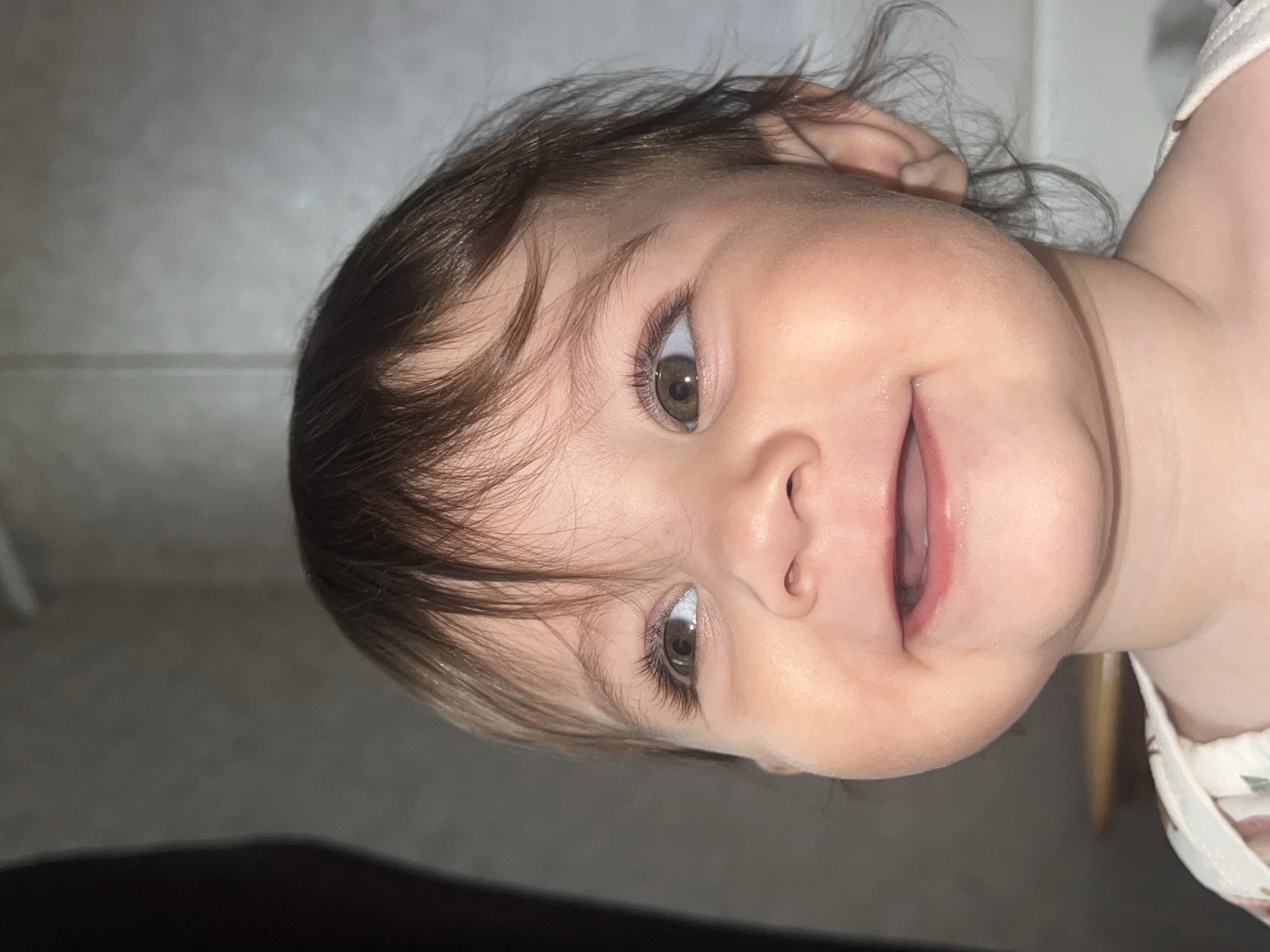 A close-up portrait shows a joyful baby, with the image rotated 90 degrees counter-clockwise. The baby has dark brown hair, styled with bangs across the forehead and some strands around the ears. Large, expressive dark brown eyes look directly at the viewer. A wide, open-mouthed smile reveals gums and suggests a happy, curious, or playful mood.
The baby's skin is fair and smooth. A small portion of a light-colored garment, possibly adorned with a subtle floral or patterned design, is visible on the right side of the frame. The background is a plain, light grey tone, suggesting an indoor setting without distinct features. The lighting is soft and appears to illuminate the baby's face evenly. No specific objects or text are visible in the background or foreground, besides the baby. The general atmosphere is one of innocence and contentment. Although the location context is Elena, Bulgaria, there are no specific visual cues within the image itself to confirm this.