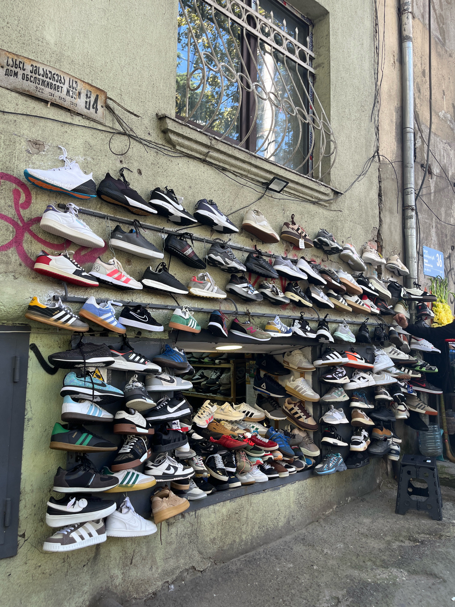 The image depicts an outdoor shoe store display on a building facade in Tbilisi, Georgia. Numerous pairs of athletic shoes are mounted on the wall, arranged in rows and sections. The shoes vary in style, color, and brand, including what appear to be sneakers and running shoes. Some shoes have visible brand logos such as Adidas and Nike. The wall itself is painted a light green, with some graffiti present near the upper left. Above the shoes, a window with a decorative metal grate is visible. A street sign with Georgian script and numbers is mounted on the wall. The ground in front of the display is concrete, with some debris and a drain visible. A foldable black stool is placed on the ground to the right. A person's hand, wearing dark clothing, is visible on the far right, interacting with the displayed items. The overall environment suggests a commercial street in an urban setting.