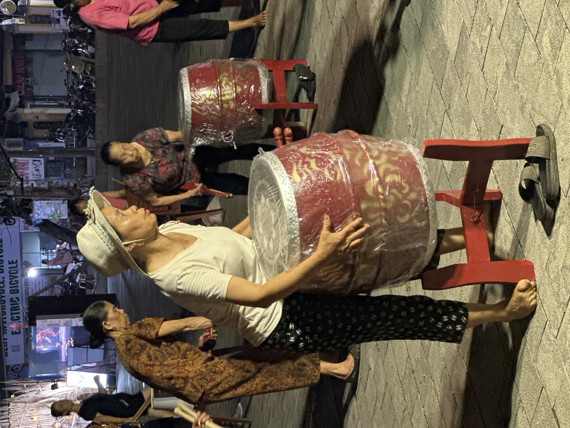 The image depicts a street scene at night in Nam Hoa Lư Ward, Vietnam. The foreground features two large, red drums, each placed on a wooden stand. The drums are wrapped in clear plastic, revealing decorative golden patterns. A woman in a white t-shirt and patterned black pants is standing next to one of the drums, with her hand resting on it. She is wearing a straw hat and appears to be preparing to play. To her left, another woman in a patterned brown dress is holding drumsticks. In the background, several other women are visible, some in pink and patterned shirts. The street is paved with interlocking stones. Behind the people, there are buildings with signs, including one that reads "ELECTRIC BICYCLE RENTAL." Motorcycles are parked in front of the buildings. The lighting suggests it is evening or night, with artificial lights illuminating the scene.