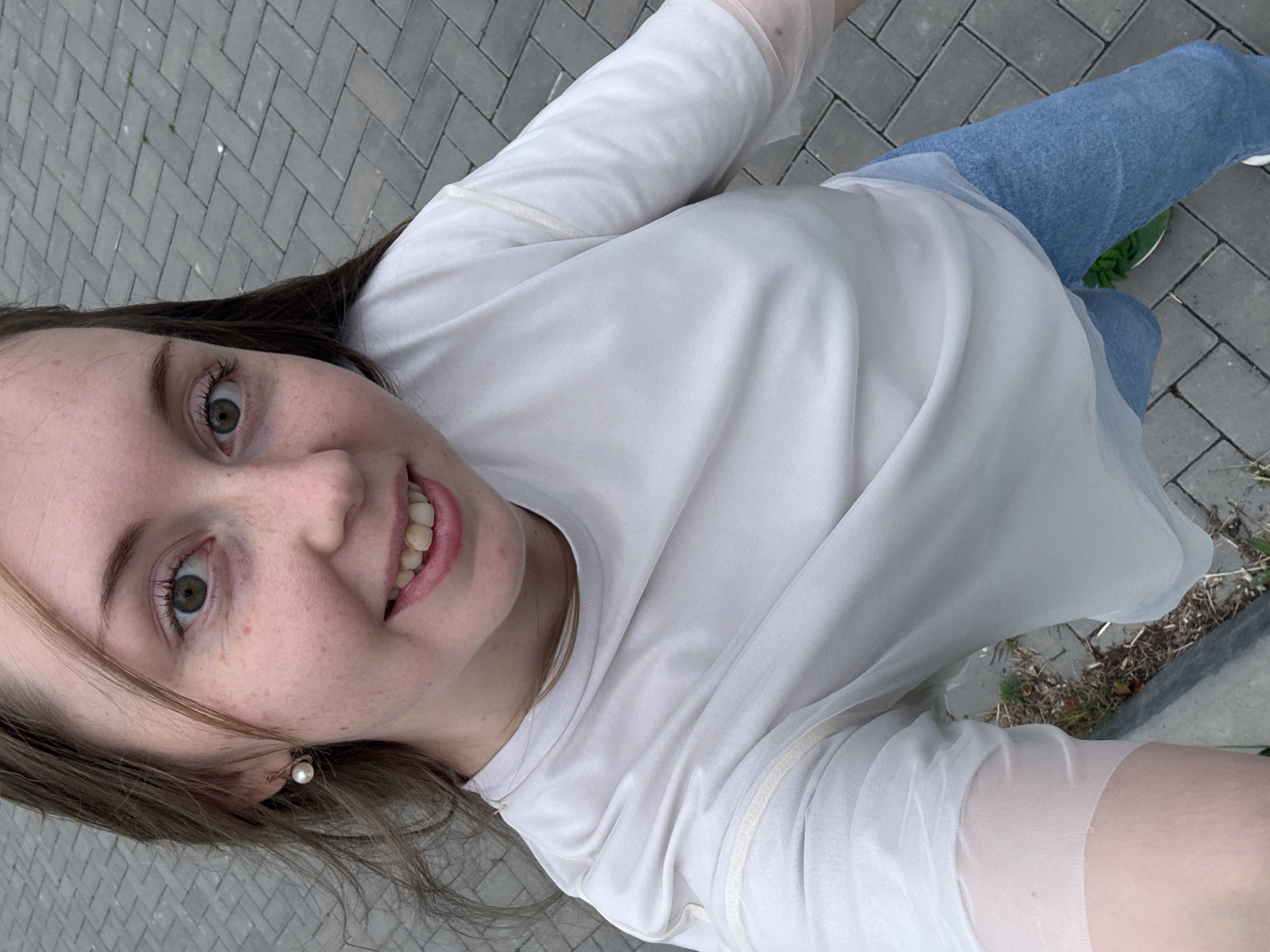 A young woman with light brown hair and green eyes is captured in a high-angle, selfie-style shot. She is wearing a white t-shirt with sheer sleeves and blue jeans. The background consists of a grey brick sidewalk. She has a pearl earring in her left ear and freckles across her nose and cheeks. Her mouth is slightly open, revealing her teeth, and she is smiling. The lighting suggests it is daytime.