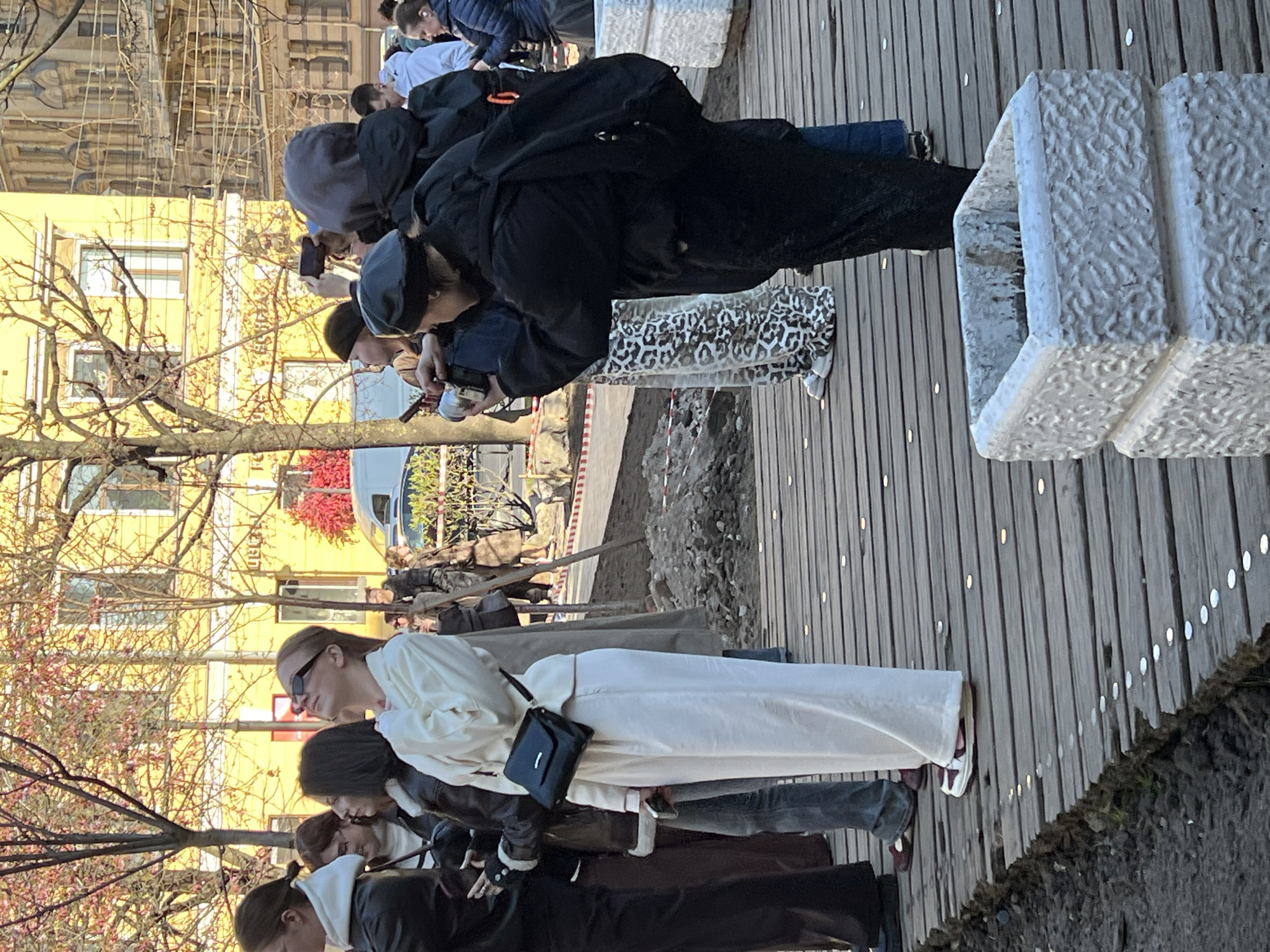 A group of people, mostly young adults, are gathered outdoors on a sunny day in Saint Petersburg, Russia. They are standing on a wooden boardwalk composed of planks with small lights embedded along the edges. Next to the boardwalk, there's a strip of dirt and gravel, and several white, textured stone blocks that appear to be seating or decorative elements.
Many individuals in the foreground and mid-ground are looking downwards or slightly forward, with several holding smartphones or small cameras, suggesting they are observing something of interest or taking pictures. One person in a black jacket and leopard print skirt is bent forward, focused on an object or event in front of them. Another individual in a white outfit and sunglasses stands further back, also looking towards the ground.
In the background, there are buildings, including a prominent yellow structure with windows. Trees with bare branches and some early spring blossoms in pink and red hues are visible, indicating it's likely springtime. A white van is partially visible further in the background. The lighting suggests it's daytime, possibly late morning or afternoon, with bright sunlight casting subtle shadows. The overall atmosphere is casual and attentive, with people gathered in a public urban space. No visible text is present in the image.