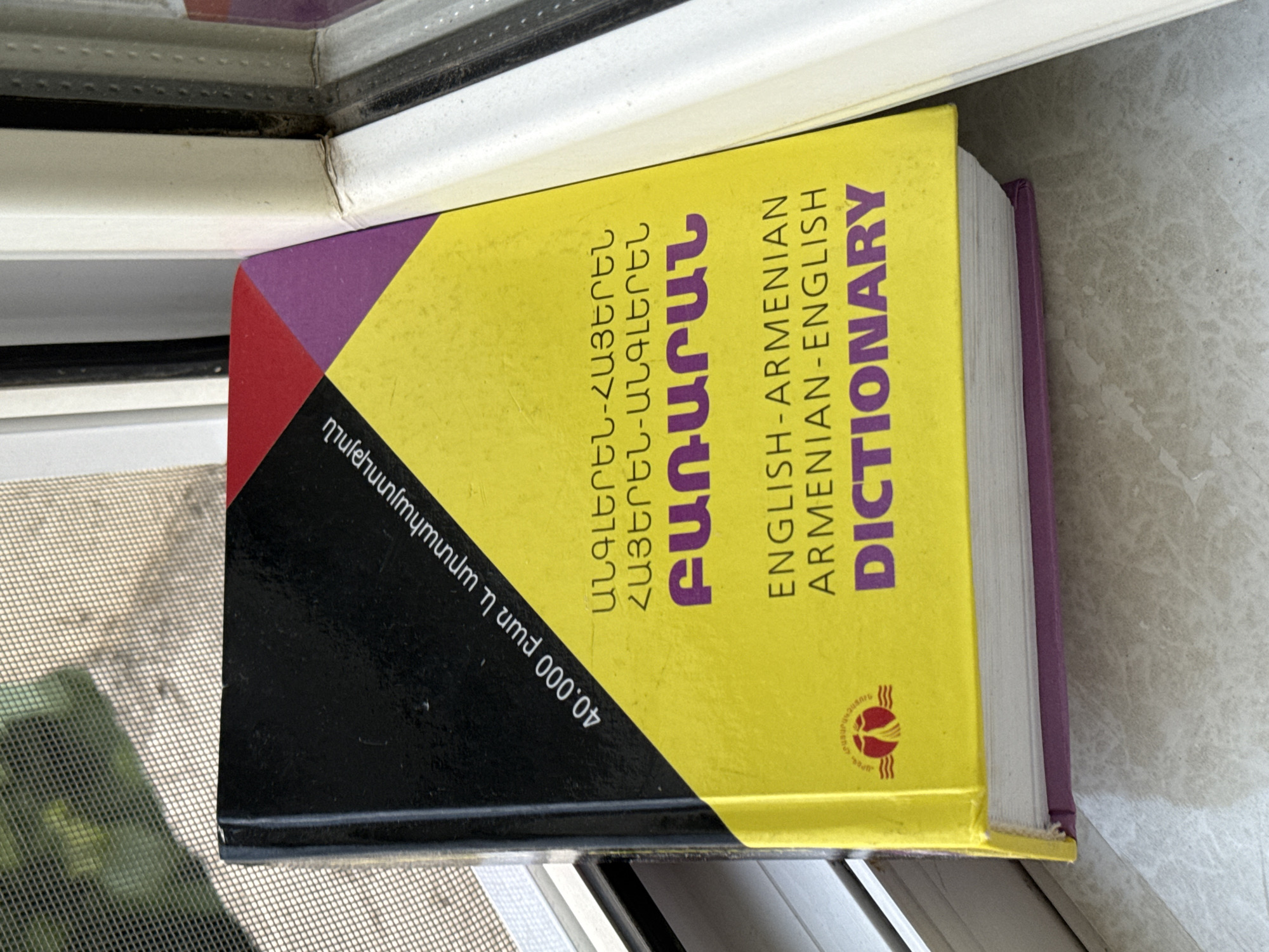 A thick, hardcover dictionary is positioned vertically on what appears to be a window sill, partially obstructing the view outside. The book's spine faces the viewer, and it is slightly tilted, leaning against the window frame.
The dictionary cover features a design with geometric sections: a large yellow area dominates, with a black section at the bottom left corner and smaller red and purple triangular shapes meeting at the center. The main title, "DICTIONARY," is prominently displayed in purple capital letters on the yellow section, along with "ENGLISH-ARMENIAN" and "ARMENIAN-ENGLISH." Additional text in Armenian script, "ԲԱՌԱՐԱՆ" (Bararan - Dictionary), "ԱՆԳԼԵՐԵՆ-ՀԱՅԵՐԵՆ" (English-Armenian), and "ՀԱՅԵՐԵՆ-ԱՆԳԼԵՐԵՆ" (Armenian-English), is also visible. On the black section of the cover, white text indicates "40.000 բառ և արտահայտություն" (40,000 words and expressions). A small circular emblem with red and yellow colors is present on the yellow part of the cover.
The dictionary's pages are visible along its right edge, appearing thick and numerous. The window sill it rests on is light-colored, possibly concrete or a similar material, showing some texture and minor blemishes. Behind the dictionary, parts of a white and dark-colored window frame are visible. To the left, a window screen with a fine mesh pattern can be seen, and a blurred green object, possibly foliage, is faintly visible through it, suggesting an outdoor scene during daylight hours. No specific activity is taking place; the book is static.