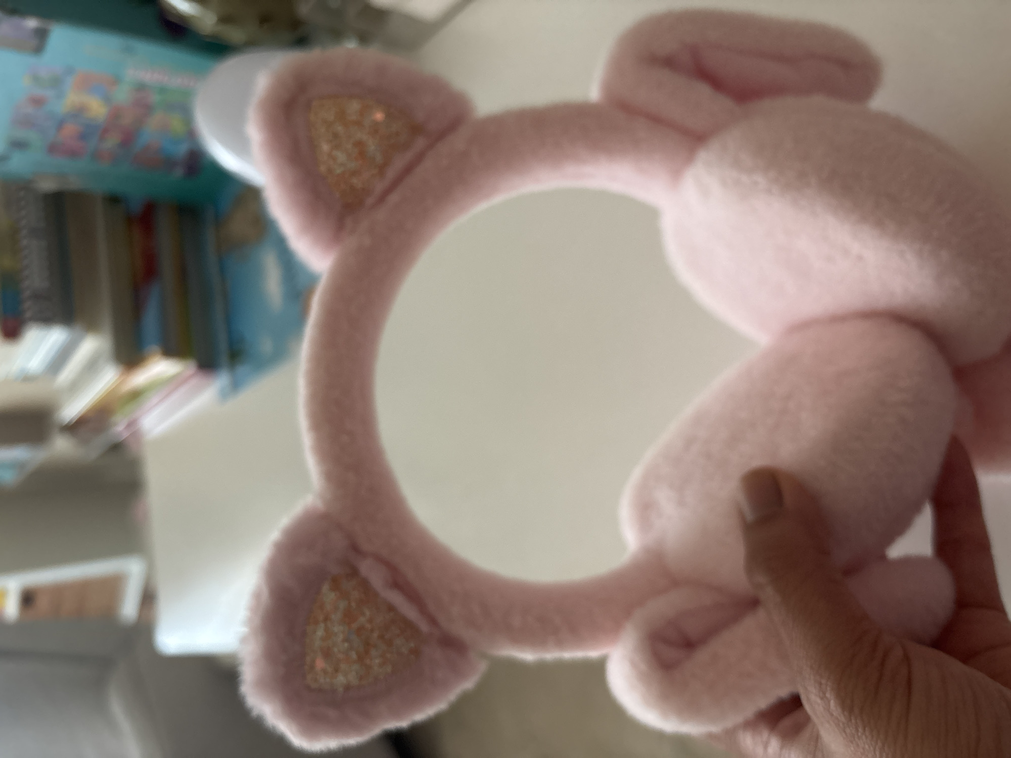A close-up, slightly out-of-focus shot shows a pair of pink plush earmuffs designed to resemble cat ears. The earmuffs have glittery pink inner ear accents. A person's hand, with a light brown complexion and short, neatly trimmed fingernails, is holding the earmuffs. The background is blurred, showing stacks of books and what appears to be a shelf unit, suggesting an indoor setting, possibly a home or office. No specific location in Dubai, United Arab Emirates, can be determined from the image.