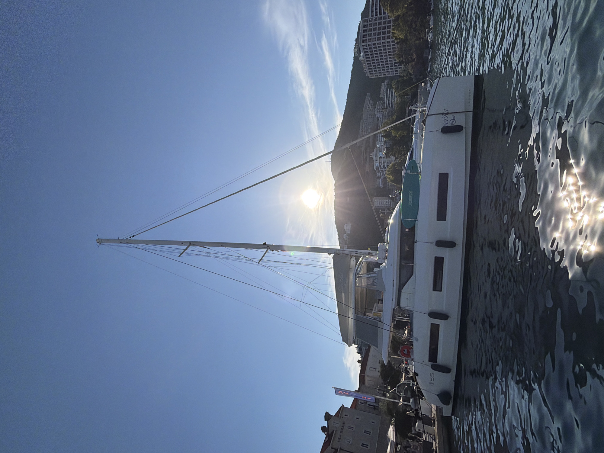 A white catamaran is docked in the harbor of Budva, Montenegro, with the sun setting behind a hilly, urbanized landscape. The catamaran, with masts and rigging visible, is positioned prominently in the foreground, its hull reflecting in the rippling water. In the background, buildings and trees are nestled into the hillside, suggesting a developed coastal area. The sky is a clear blue, transitioning to lighter hues near the horizon where the sun's rays create a bright glare. A few boats are visible further out in the water. The scene depicts a peaceful evening in a Mediterranean coastal town.