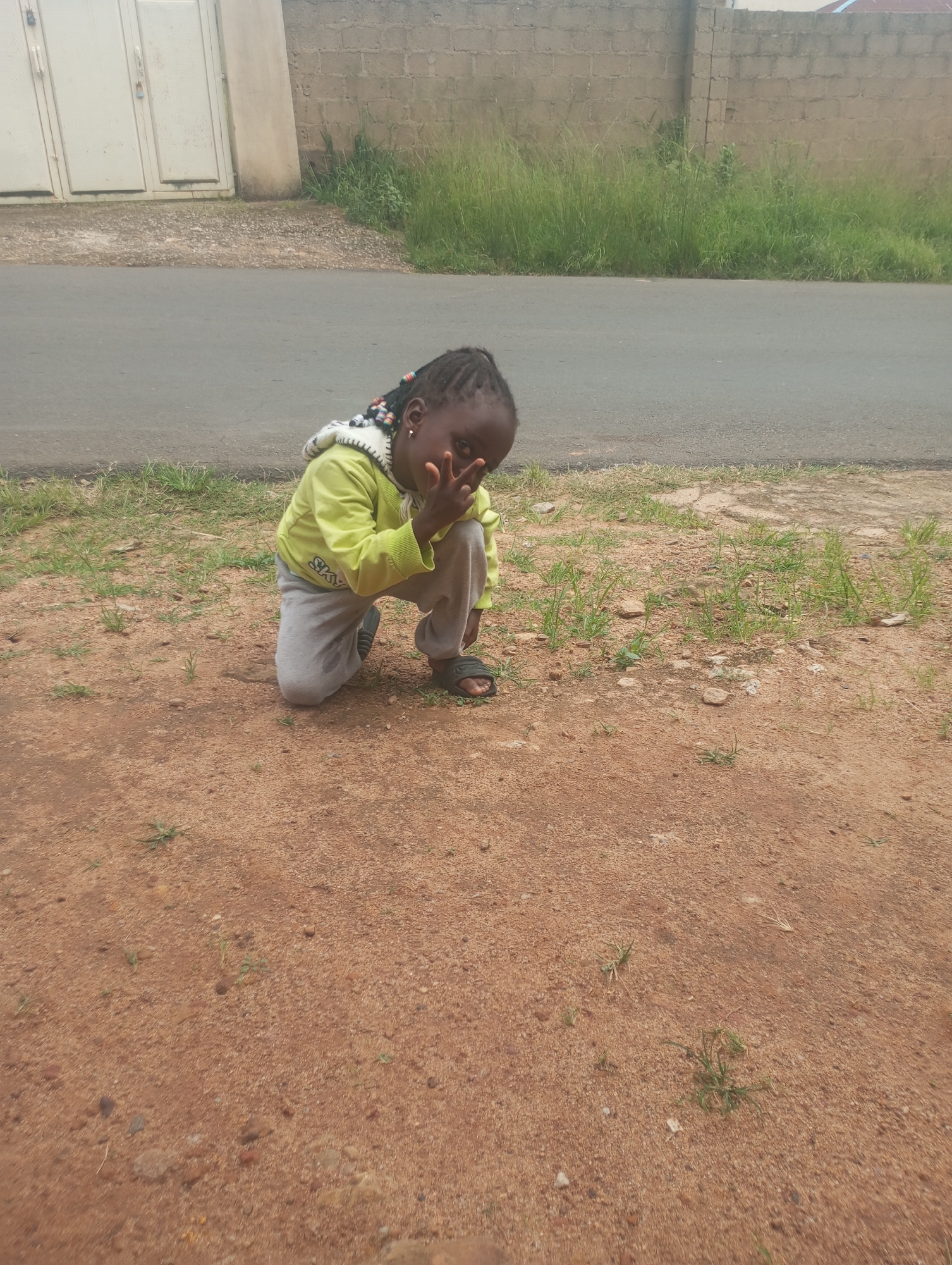 A young Black girl with braided hair adorned with beads kneels on reddish-brown dirt by a paved road. She is wearing a lime green hooded sweatshirt and gray sweatpants. Her left hand is covering her face, obscuring part of her features, and her right hand is raised with her fingers splayed. In the background, a grey wall constructed of concrete blocks runs along the edge of the road, with overgrown green grass in front of it. To the left, a large, light-colored gate with vertical panels is visible. The scene appears to be an outdoor environment in a residential or street area, potentially in the city of Jos, Nigeria, given the typical architecture and ground cover.