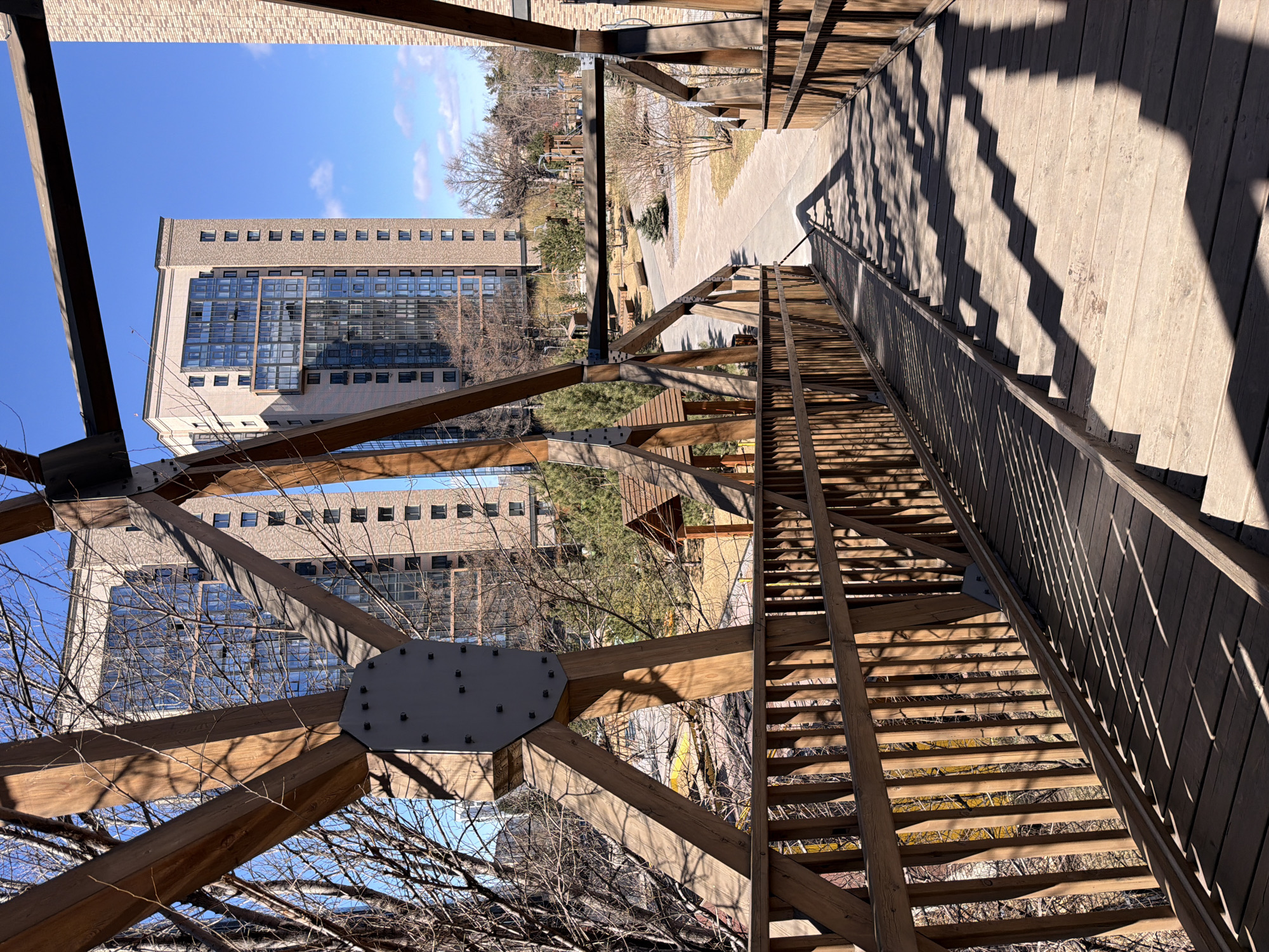 The image depicts a pedestrian bridge in Krasnoyarsk, Russia, offering a view towards a building. The bridge is constructed of wood and steel, with visible support beams and a bolted metal plate. Sunlight casts shadows across the bridge's wooden planks and handrails. In the background, a modern building with multiple floors and glass windows is visible. Bare trees surround the structure. The sky is clear and blue.
